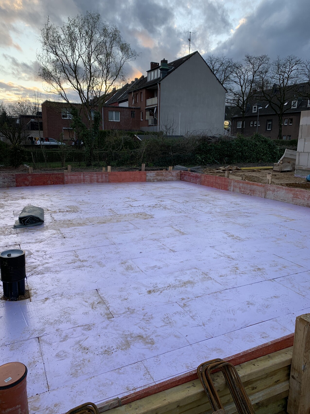 Construction site with white insulation membrane, red border, construction tools; background: houses and trees.