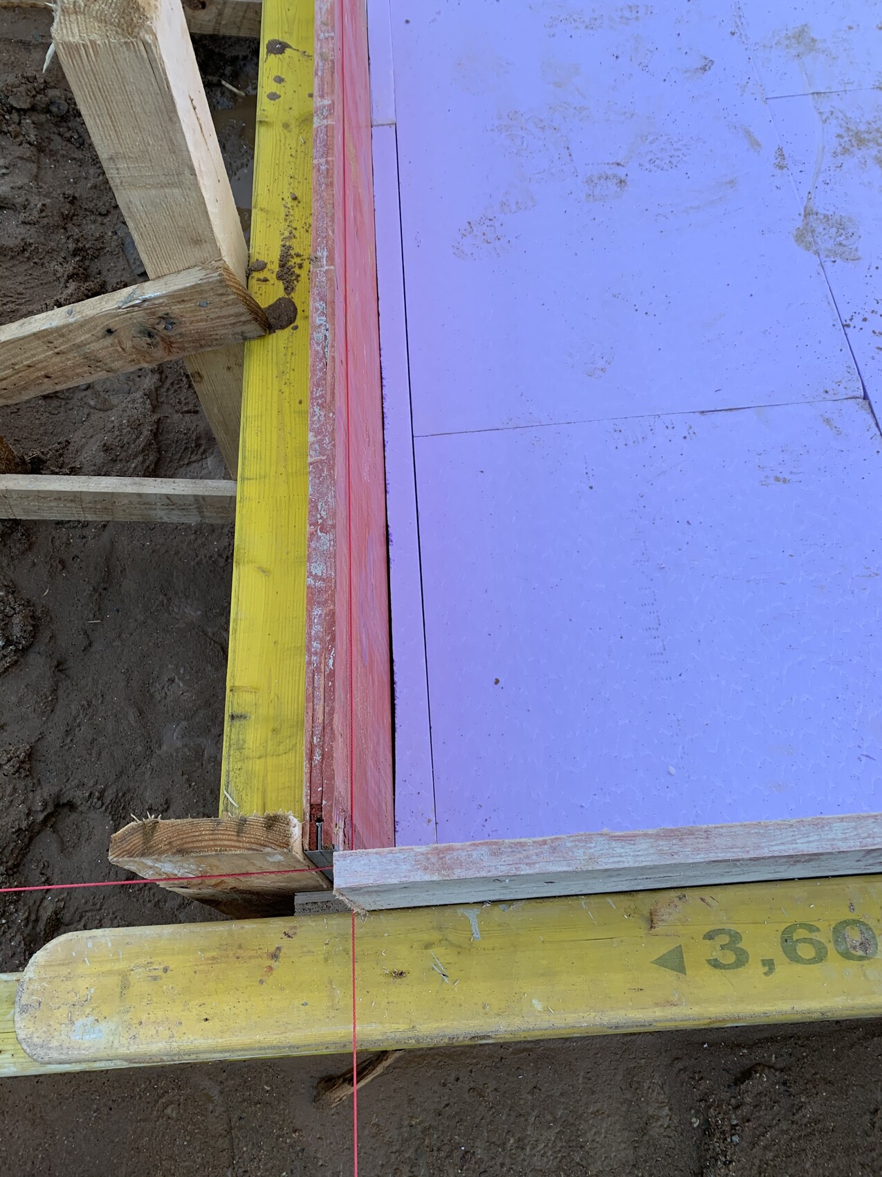 Construction site: purple insulation boards in wooden frame, red string line, yellow measuring rod on the ground, soil.