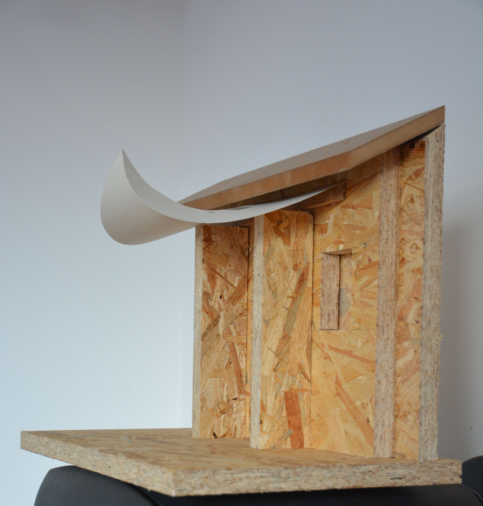 Skulptur aus OSB-Holzplatten mit einer weißen, gebogenen Blattform darauf.