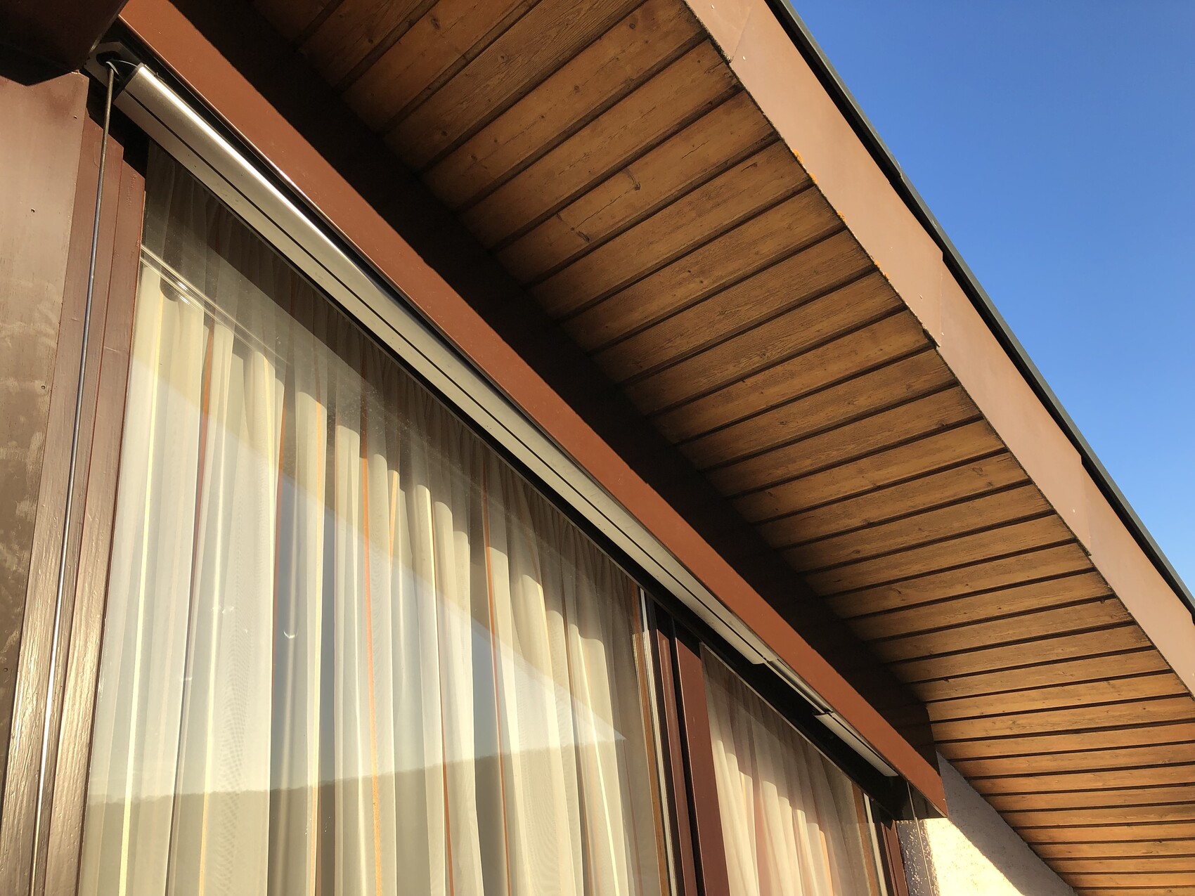 Exterior view of a wooden house with a window and curtains, overhanging wooden roof, blue sky.