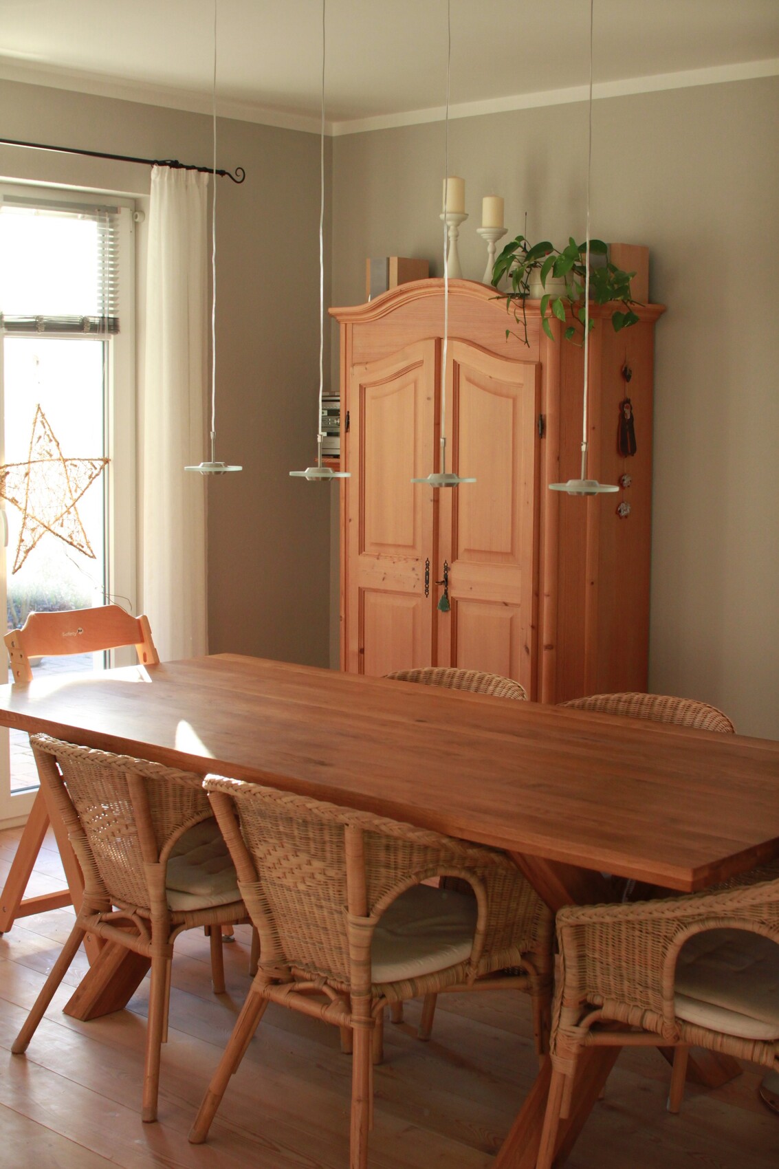 Bright dining room: wooden table with wicker chairs, large wooden cabinet, plant, and window decorations.