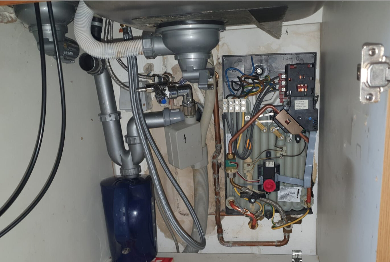 Under the kitchen sink: pipes, P-trap, hoses, and heating/water system with controls.