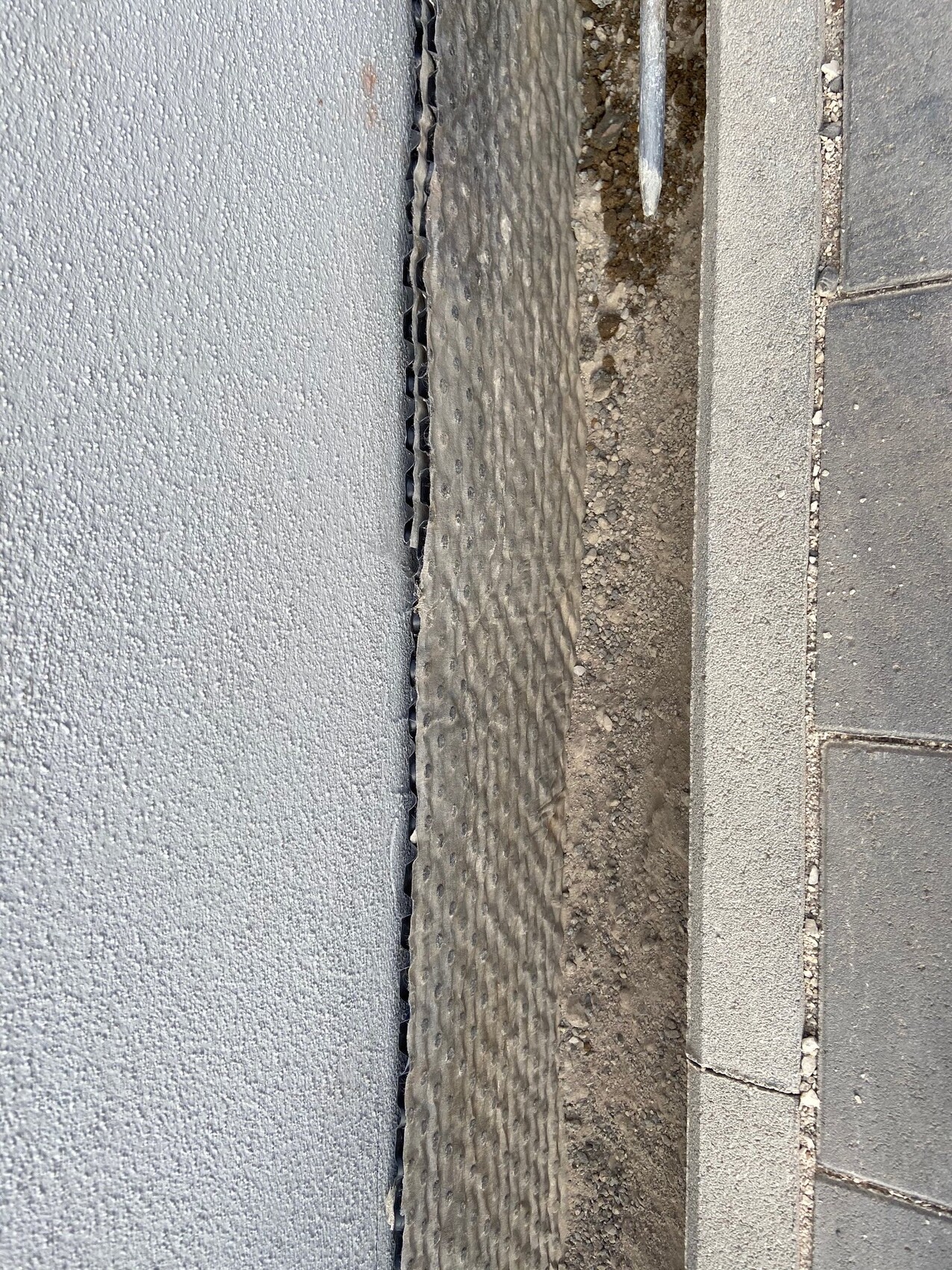 Gap between gray wall and paving edge with dark, ribbed profile and soil next to it.