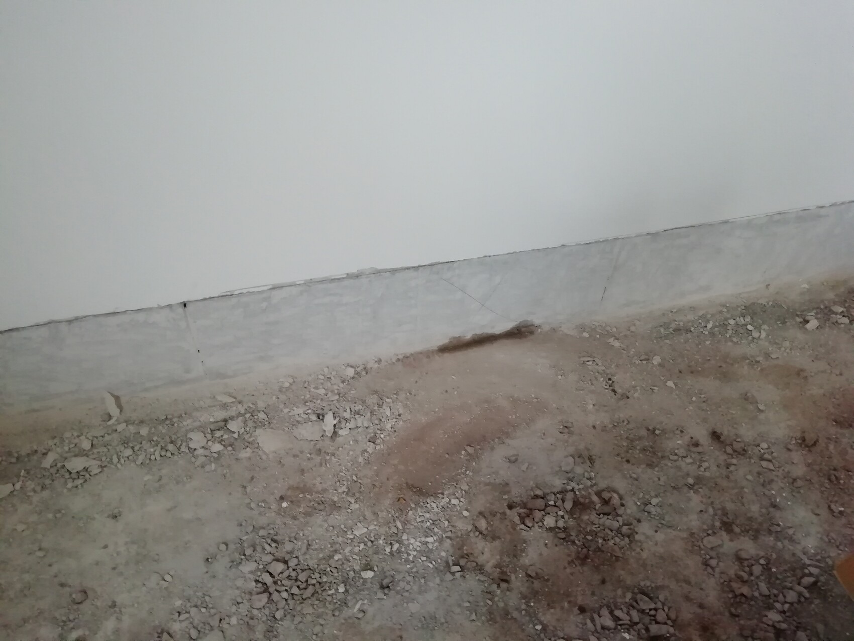 Weiße Innenwand mit grauer Betonleiste am Boden; unbefestigter, kiesiger Erdboden mit Steinen.