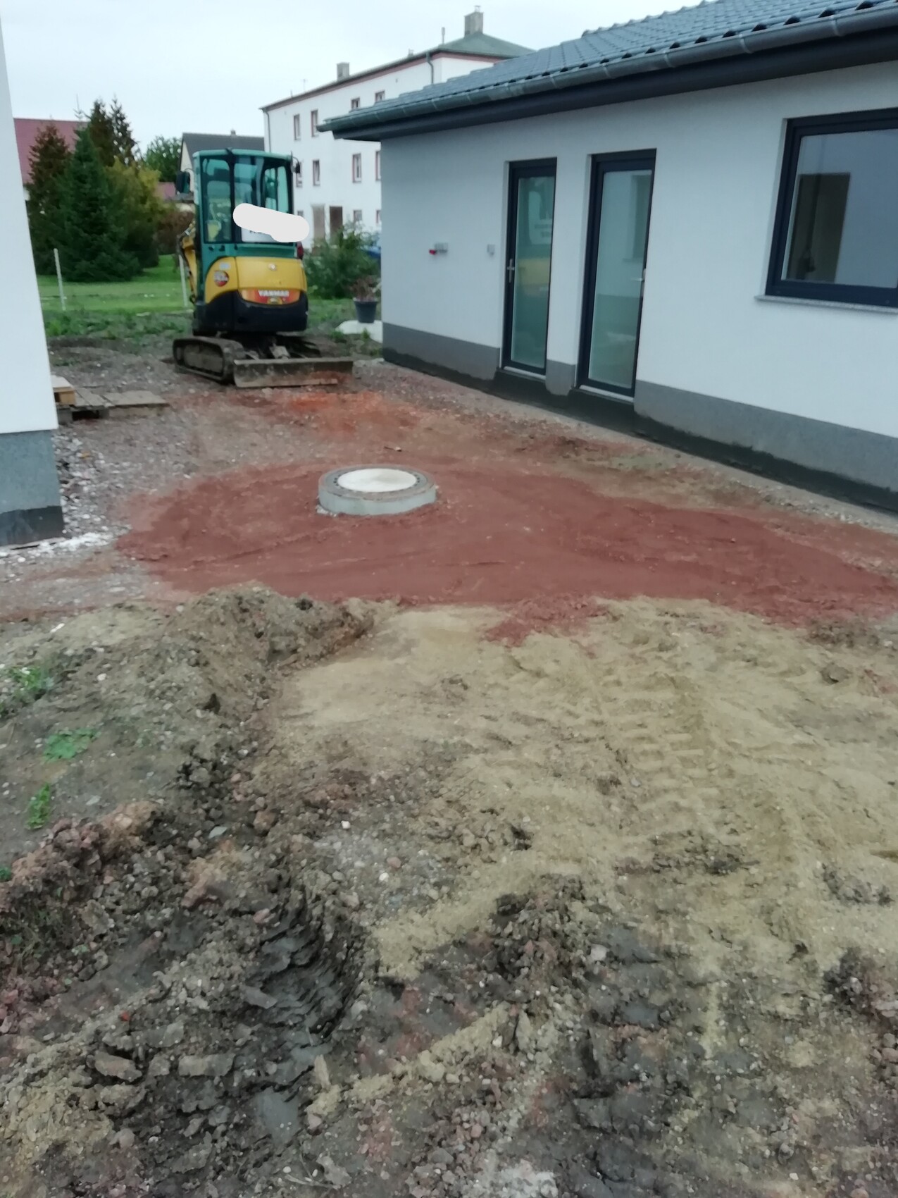 Kleiner Bagger auf Baustelle neben Wohnhaus; roter Boden mit runder Abdeckung im Erdreich