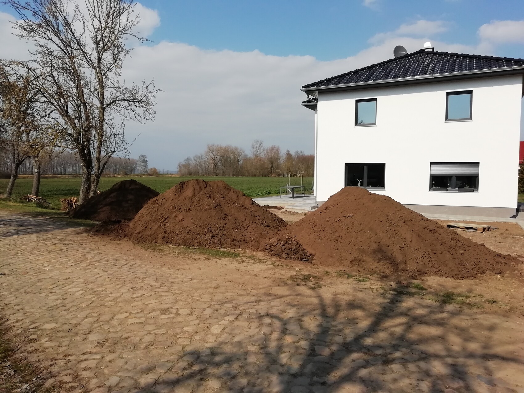 White two-story house; in front of the house, earth excavation piles, paved courtyard, row of trees.
