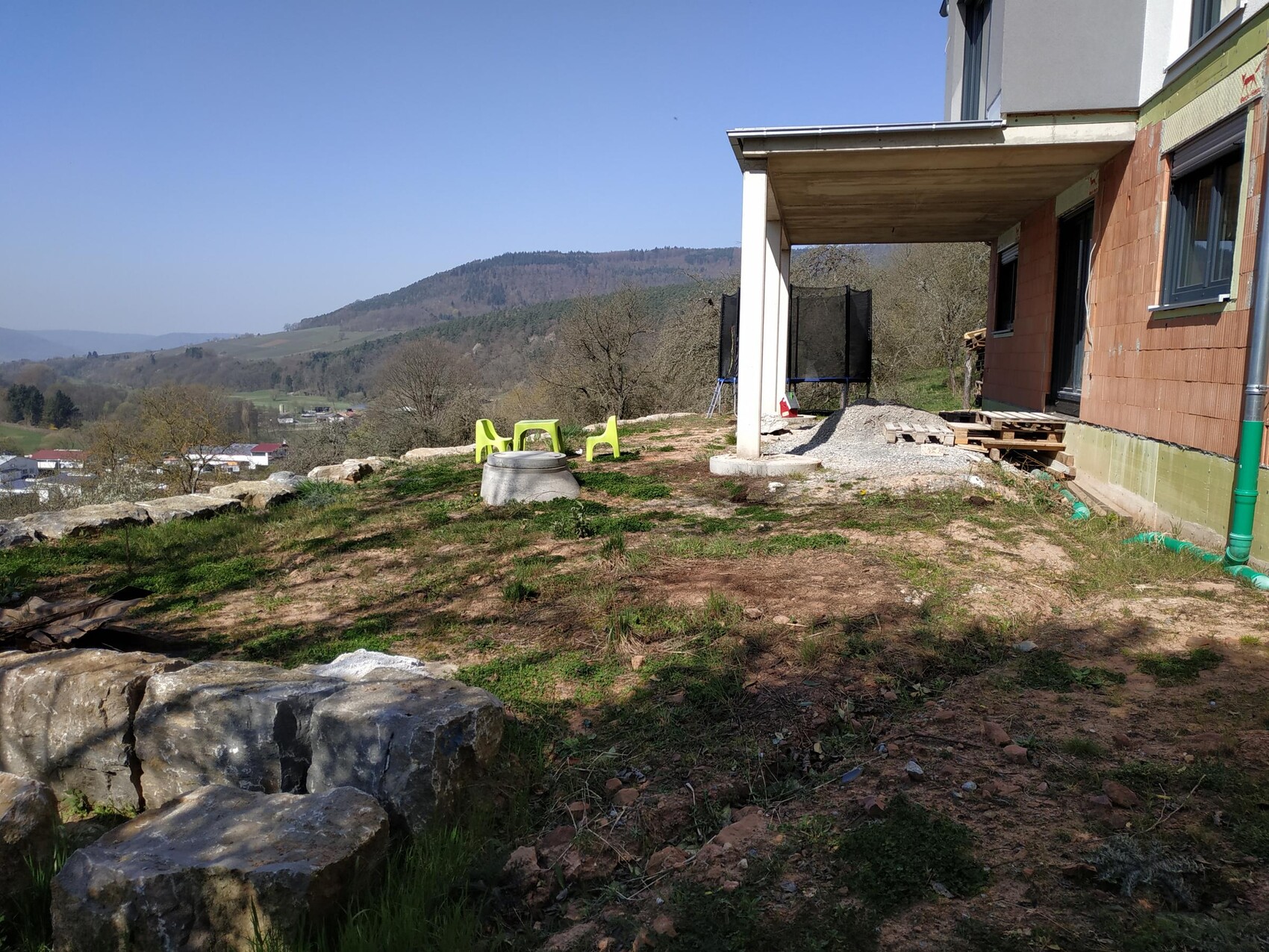 Unfinished exterior construction of a house with brick wall, pallets, trampoline and garden chairs on uneven yard