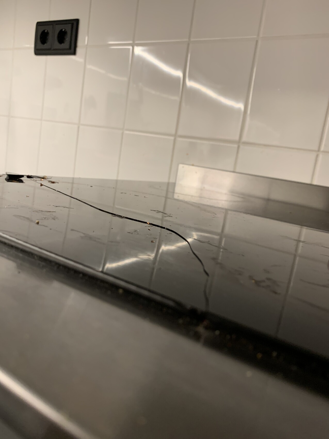 Black glass ceramic cooktop with hairline crack, reflection of white tiled wall in the background.