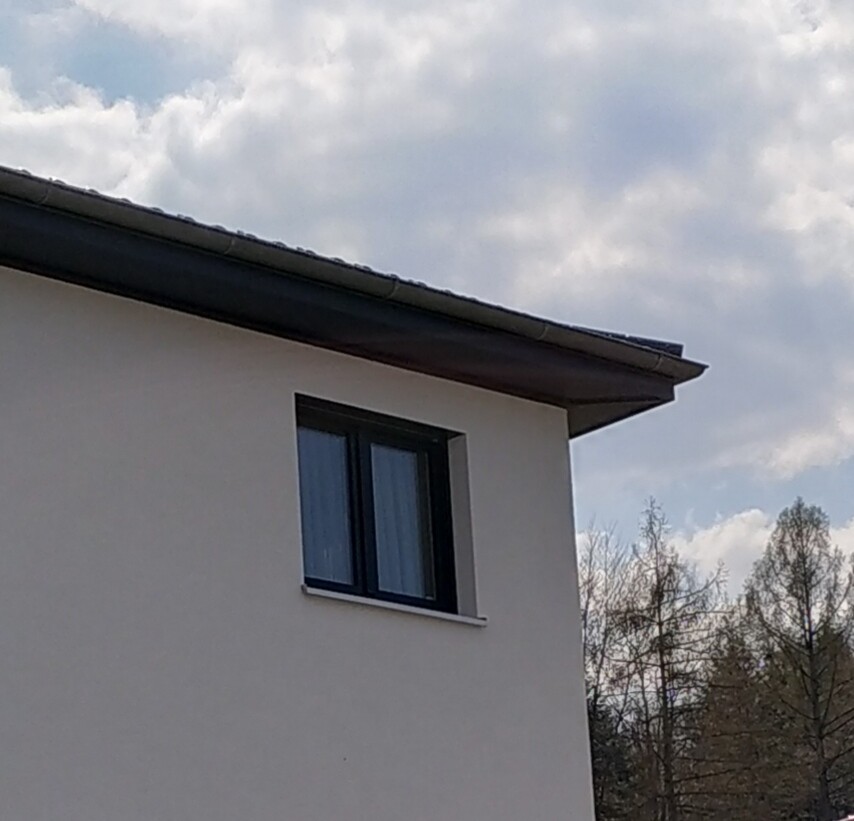 White house wall with dark-framed window, sloped roof and trees in the background.