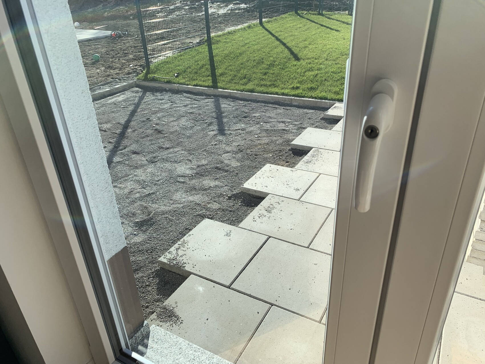 View through glass door of steps made of paving stones leading to a grass-covered garden behind a fence.