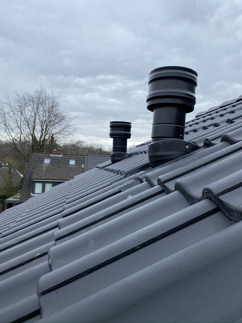 Roof with gray tiles and two black ventilation chimneys, cloudy sky.