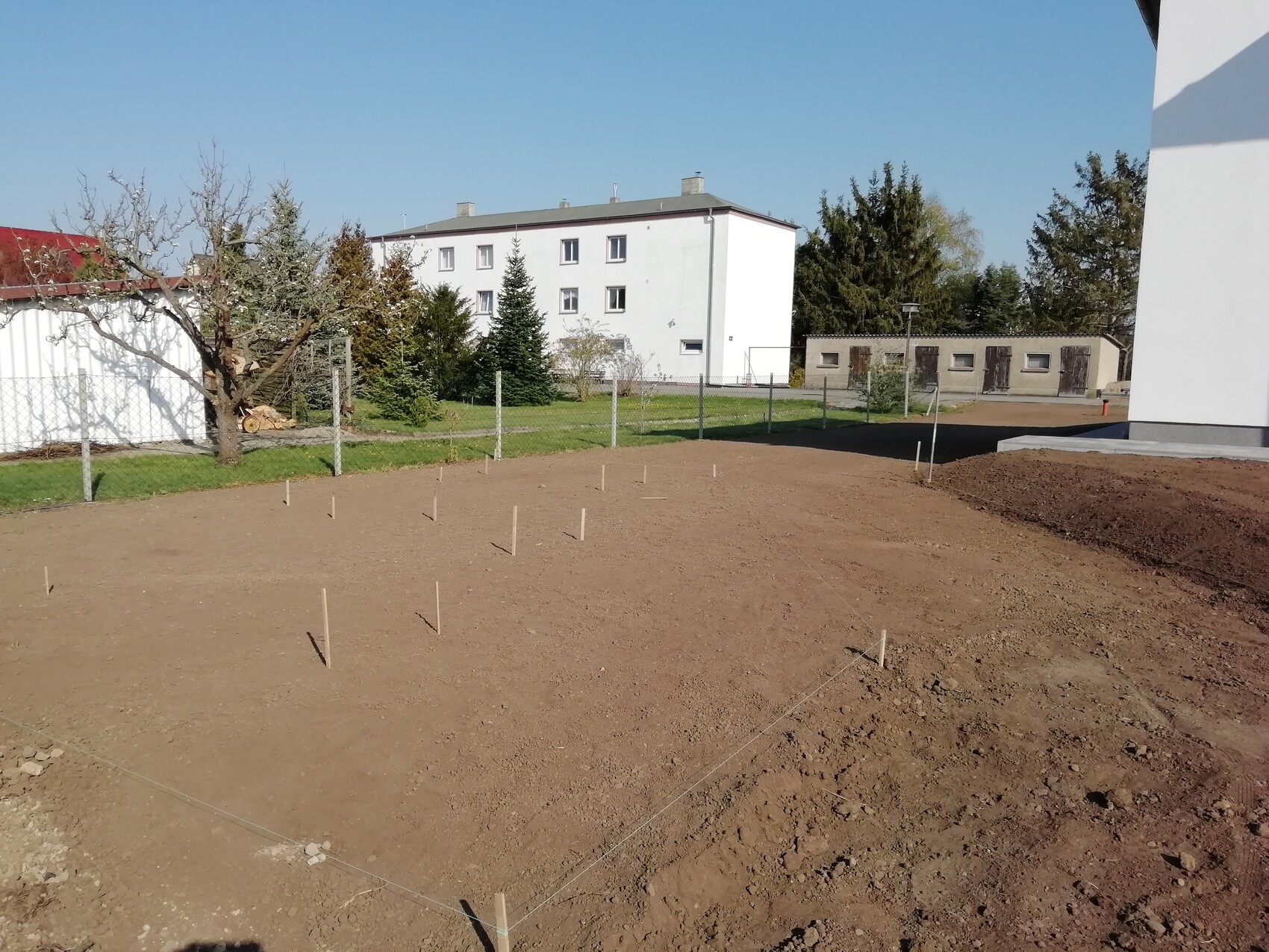 Baugrundstück mit Pfostenmarkierungen und Drahtzaun, dahinter weißes Mehrfamilienhaus und Bäume.