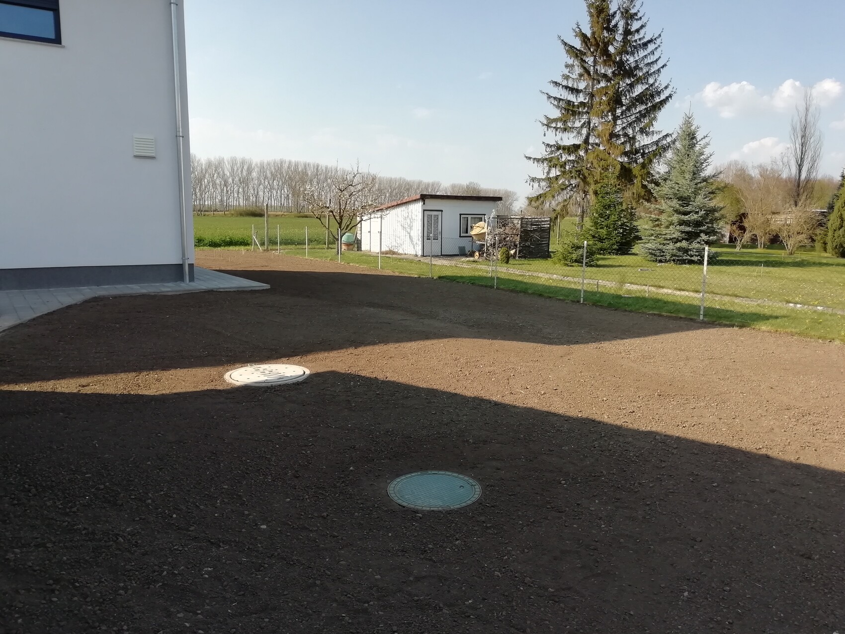 Außenbereich eines Hauses: unbefestigter Erdplatz mit zwei runden Abdeckungen, Garten hinter Zaun.