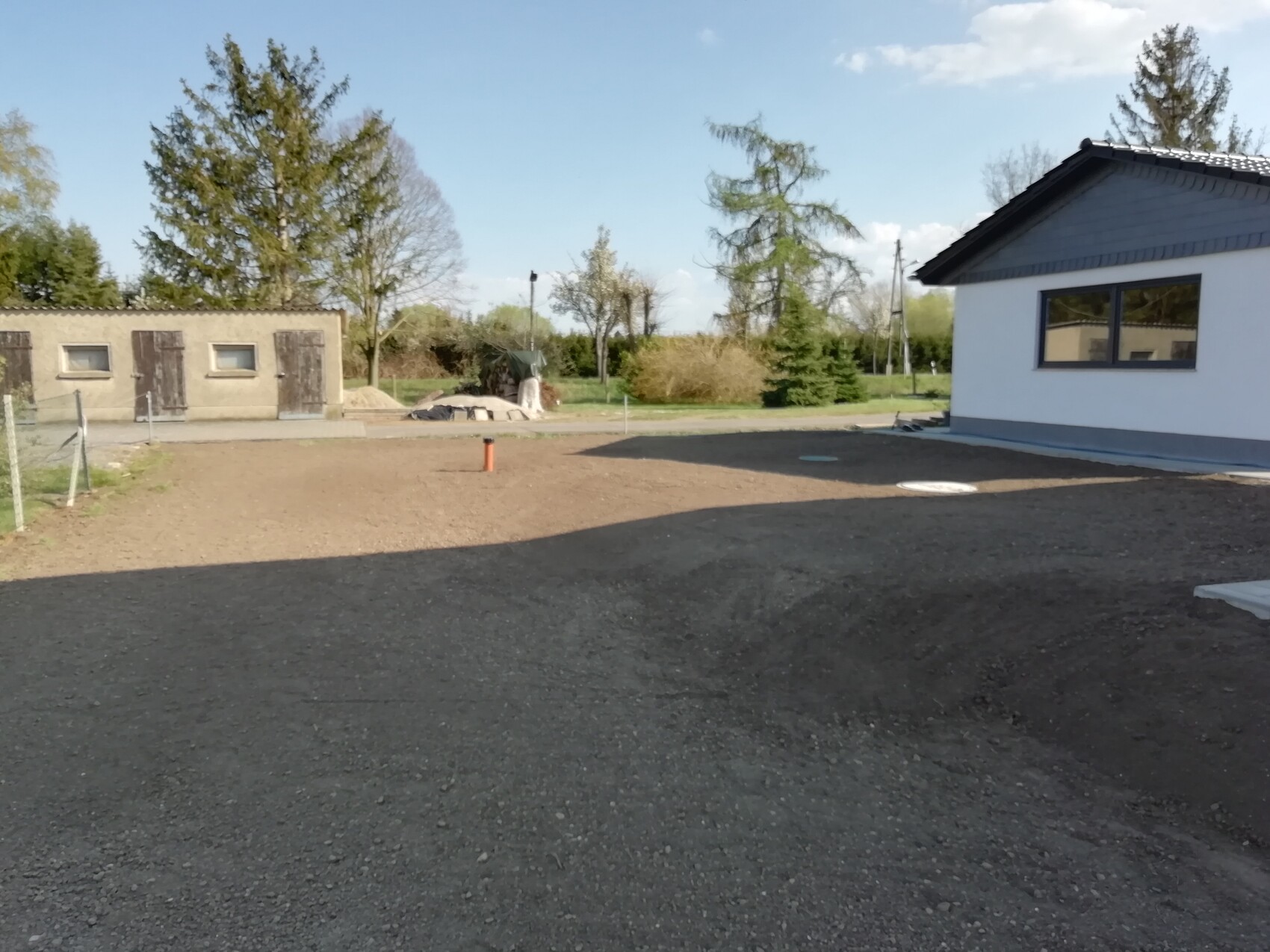 Kiesplatz vor weißem Gebäude rechts, links eine Bauhütte mit Türen, Bäume im Hintergrund.