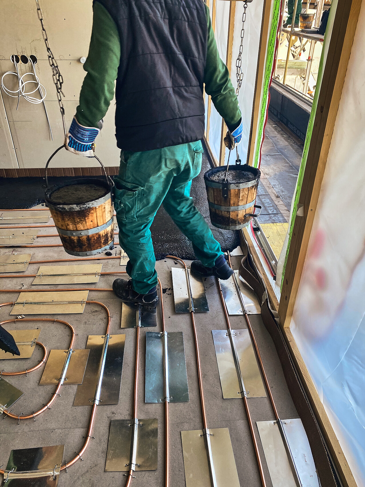 Construction worker carries two buckets of mortar; floor with copper pipes and metal plates.