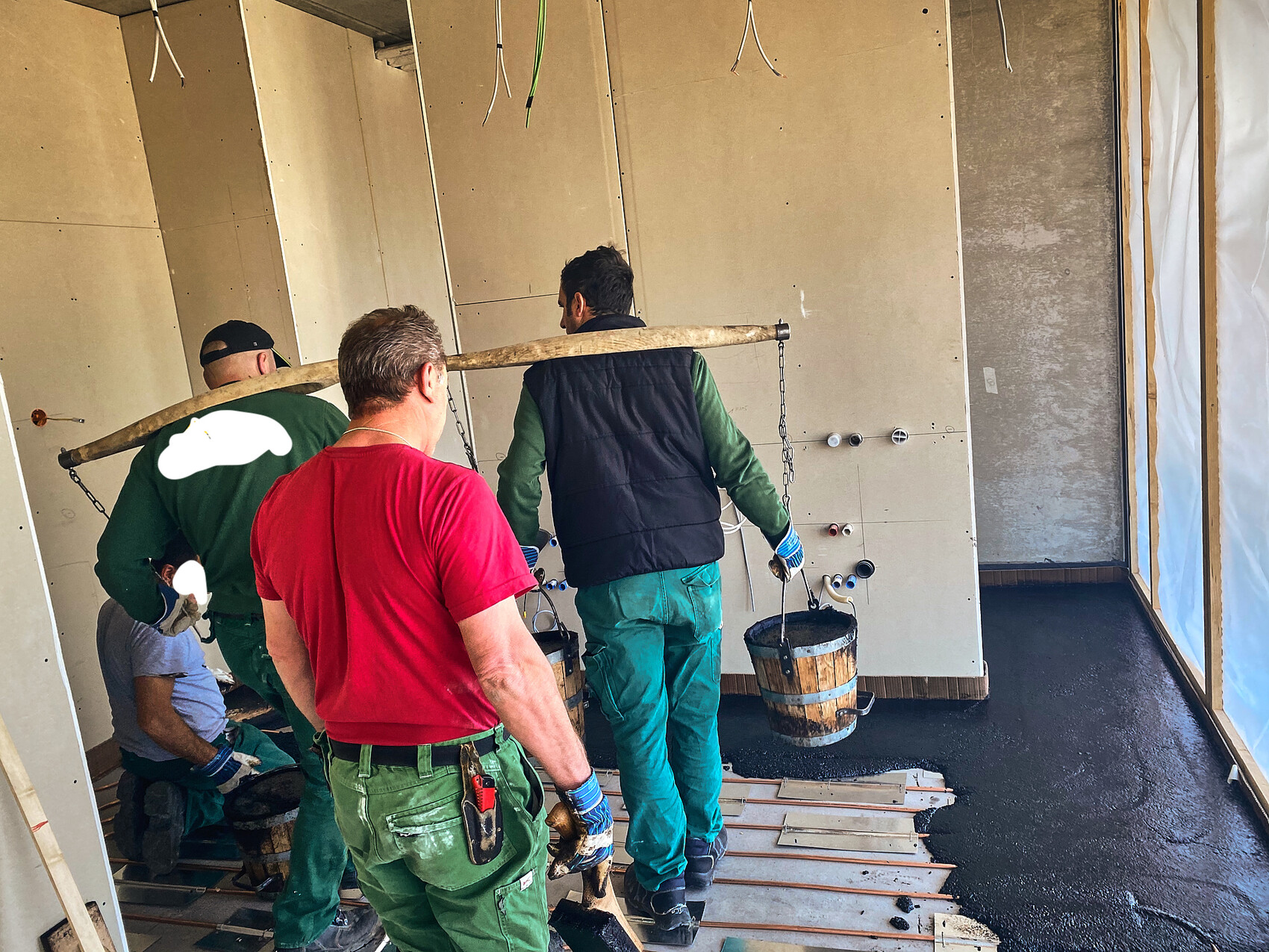 Several construction workers carry a bucket of concrete on a long wooden pole during interior construction.