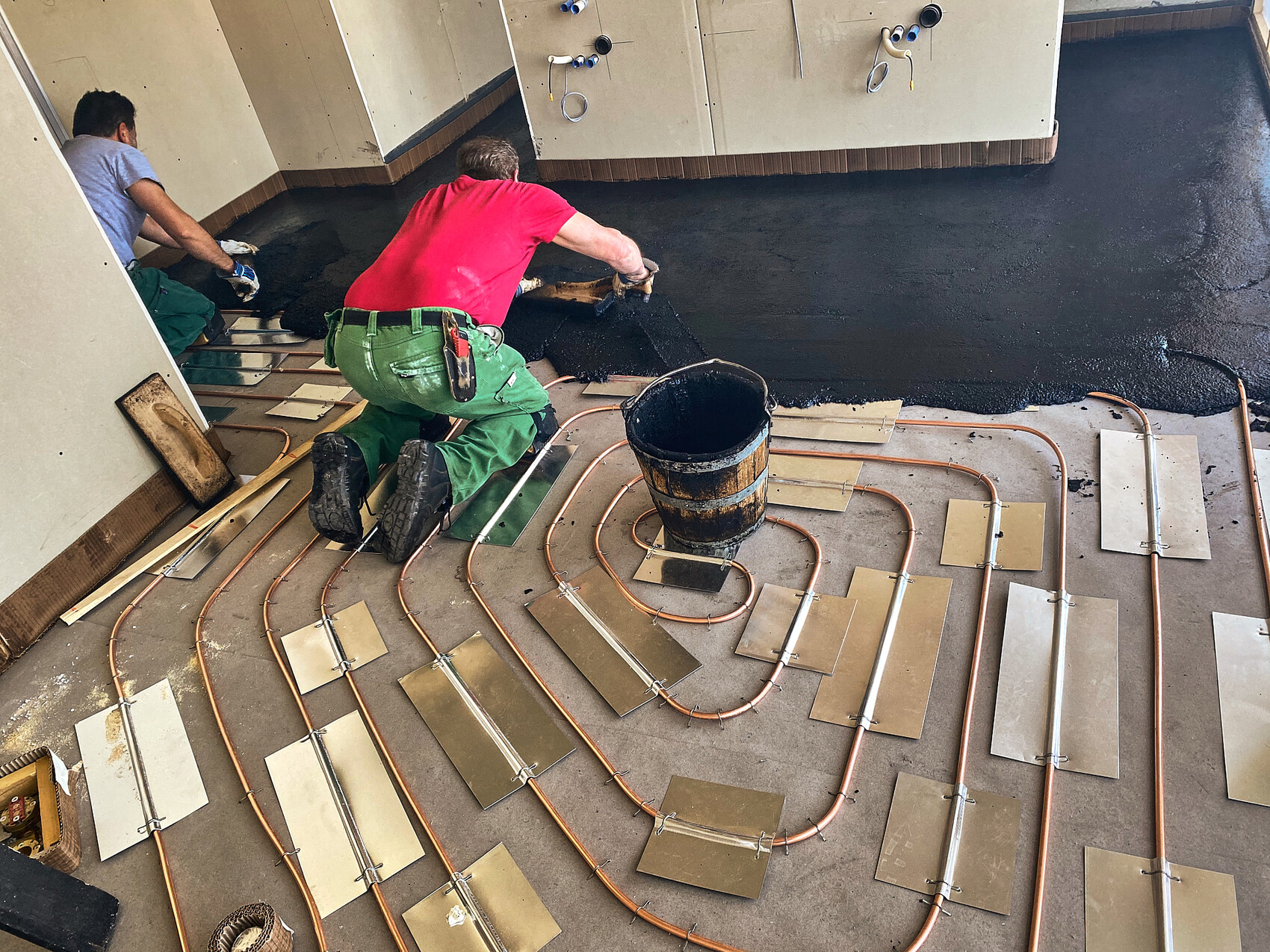 Two construction workers lay copper pipes for underfloor heating and spread black sealing compound.