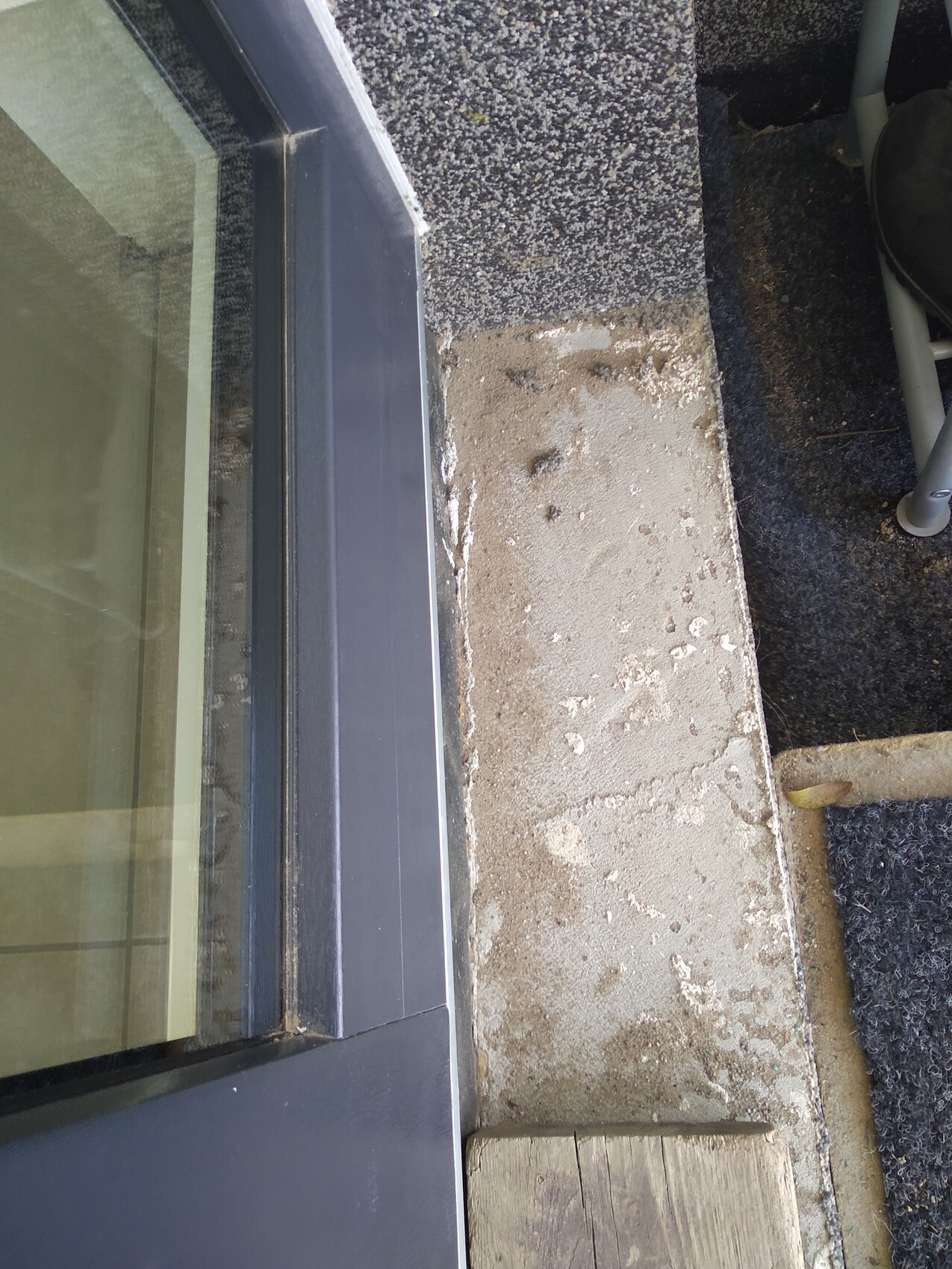 Exterior photo of a door: raw concrete step with stains, dark door frame and doormat on the right.