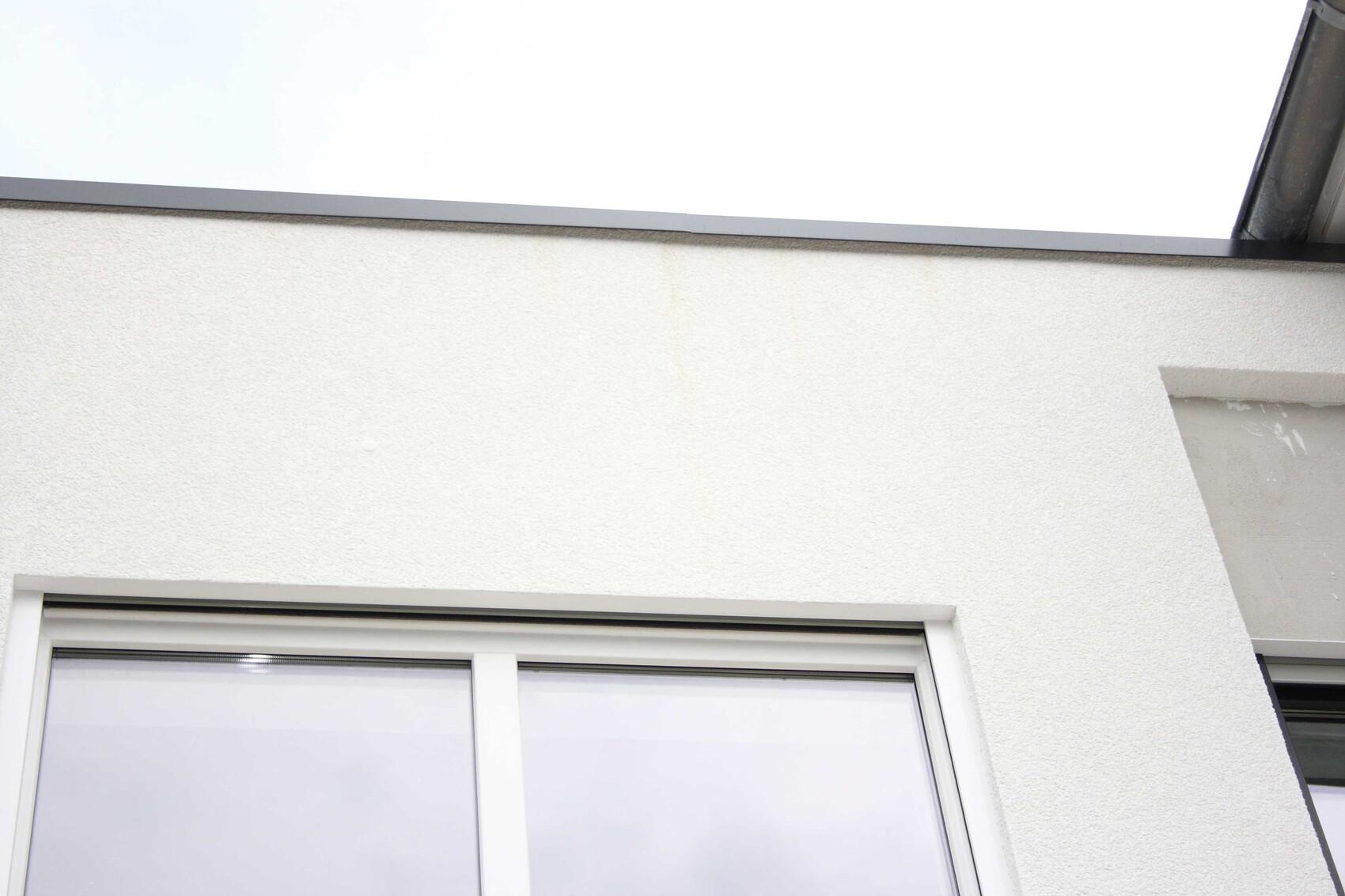 White plastered exterior wall with window at bottom left; grey edge at top, slight water stains.