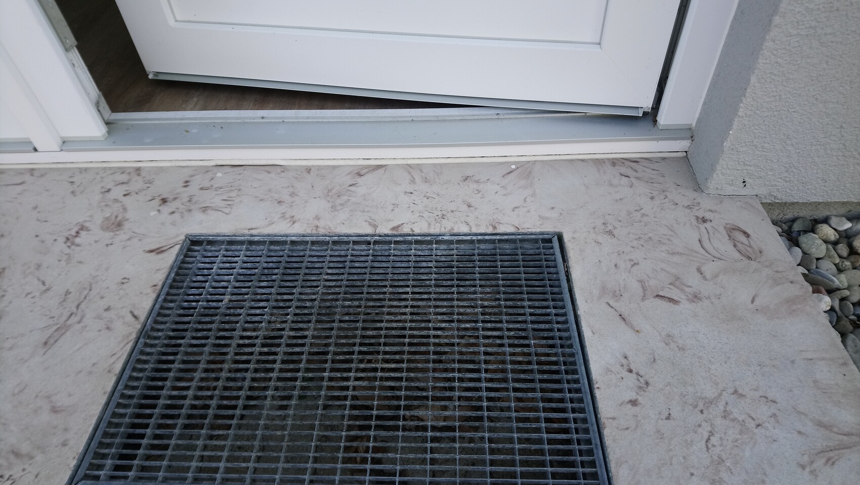 White front door slightly open; in front a metal mesh cover in light concrete, gravel on the right.