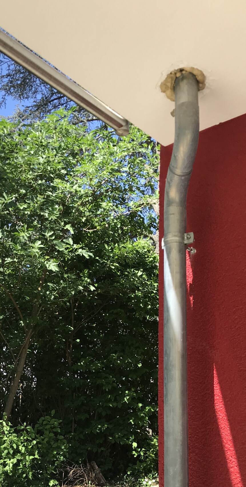 Exterior area: vertical metal downpipe on a red wall, under a white ceiling, green hedge and blue sky.