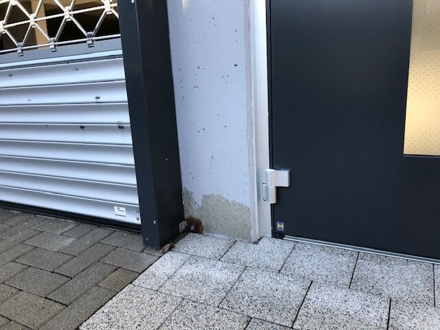 Exterior view: gray door area on the right, metal roller shutters on the left, concrete wall with rust stains at the bottom.