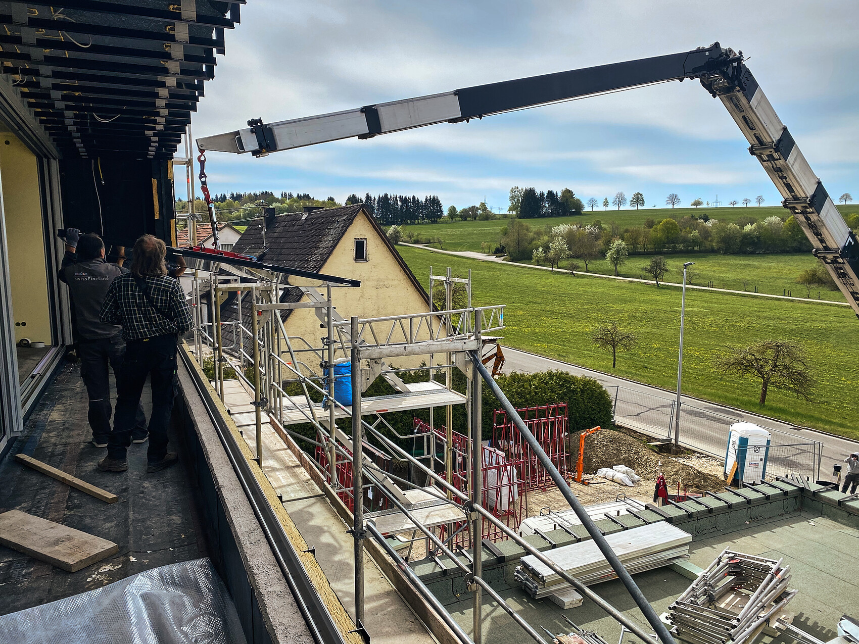 Construction site: scaffolding, crane arm, two workers at a construction wall, view of green landscape.