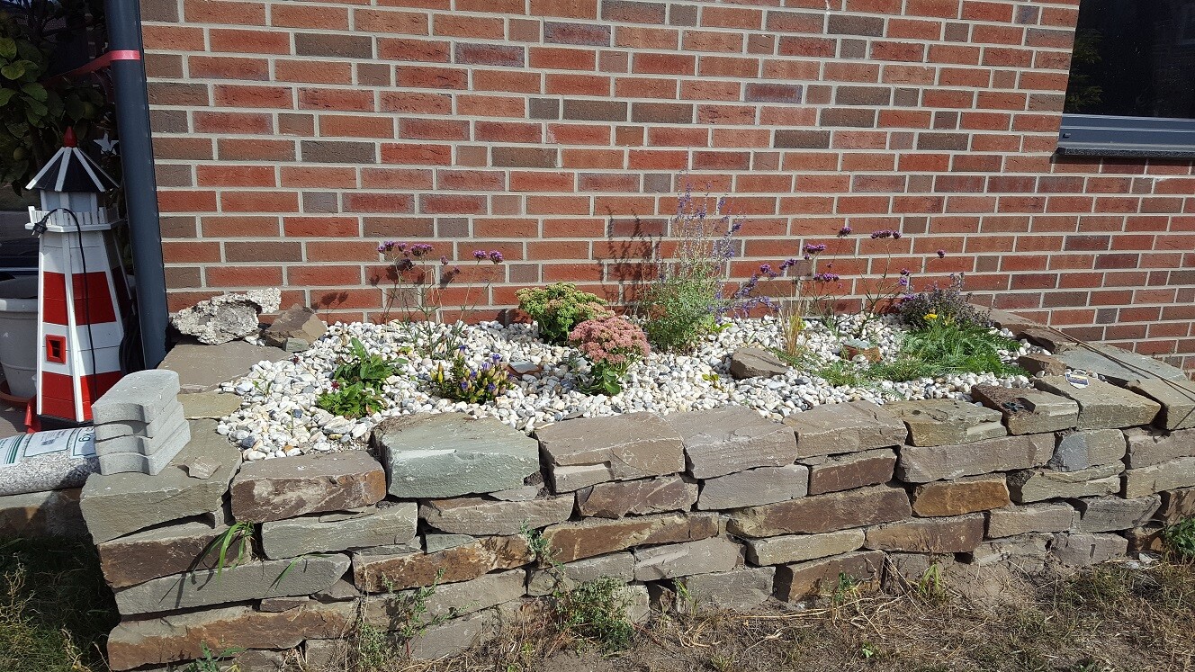 Edge of stacked stone slabs surrounds a white gravel flower bed in front of a red brick house.
