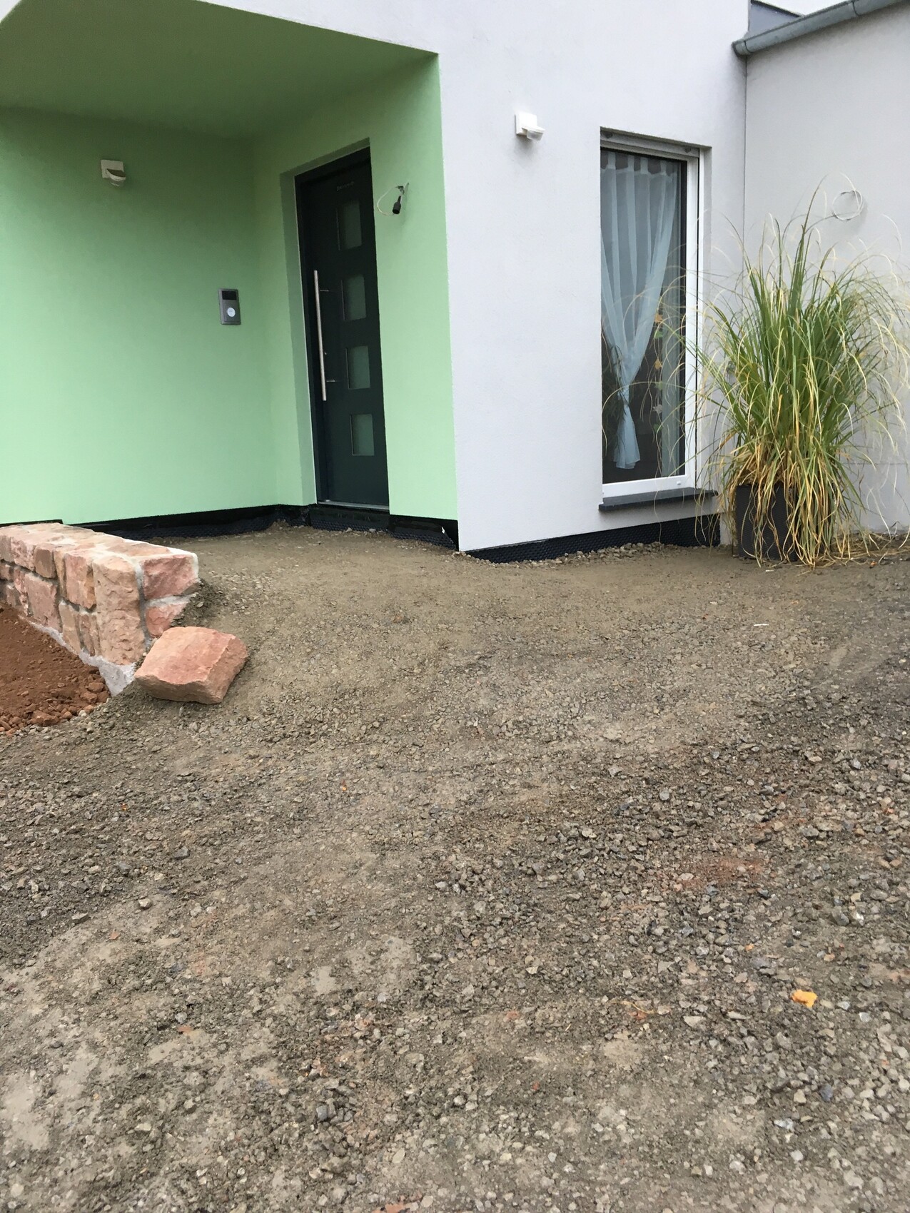 Modern front entrance: green wall, dark door, window with curtains, grasses in a planter, gravel path.