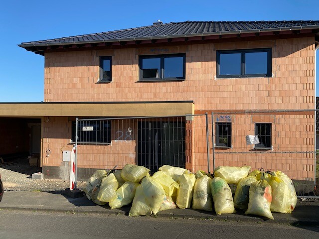 Unfinished two-story brick house with construction fence and yellow garbage bags in front of the entrance.
