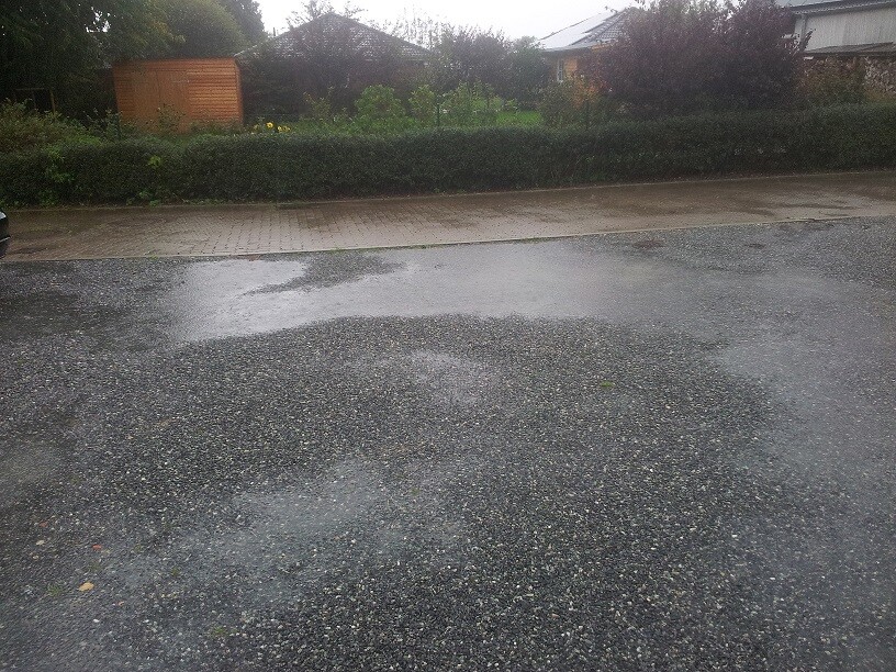 Wet forecourt with puddles in front of residential house, hedge and garden in the background