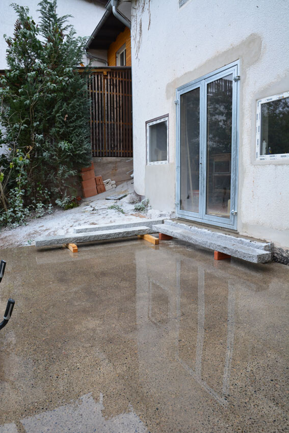 Exterior view of a house with glass sliding door; concrete bridge on wooden supports above a wet area.