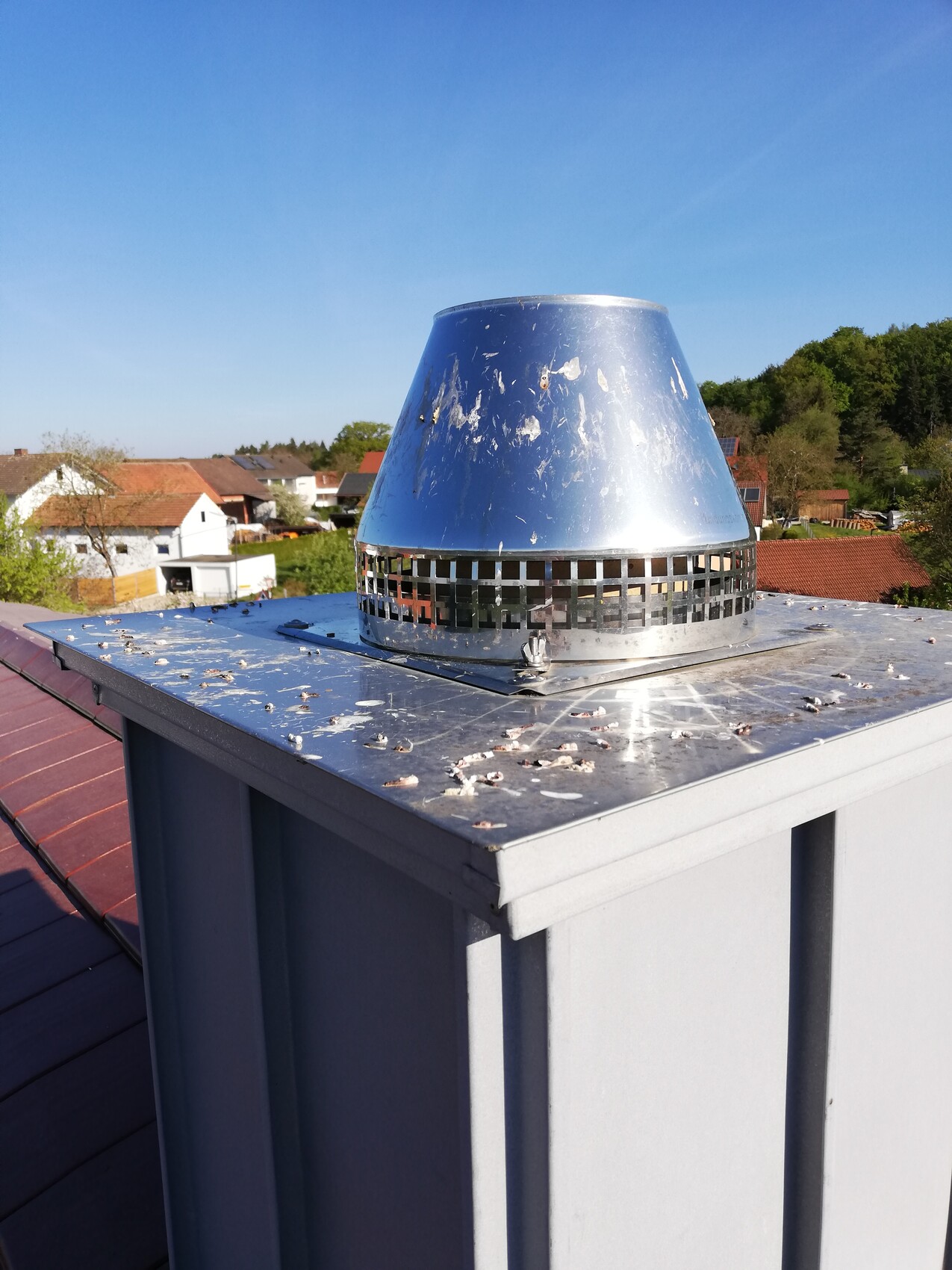 Conical metal chimney with ventilation ring on rooftop, view of houses and green surroundings.