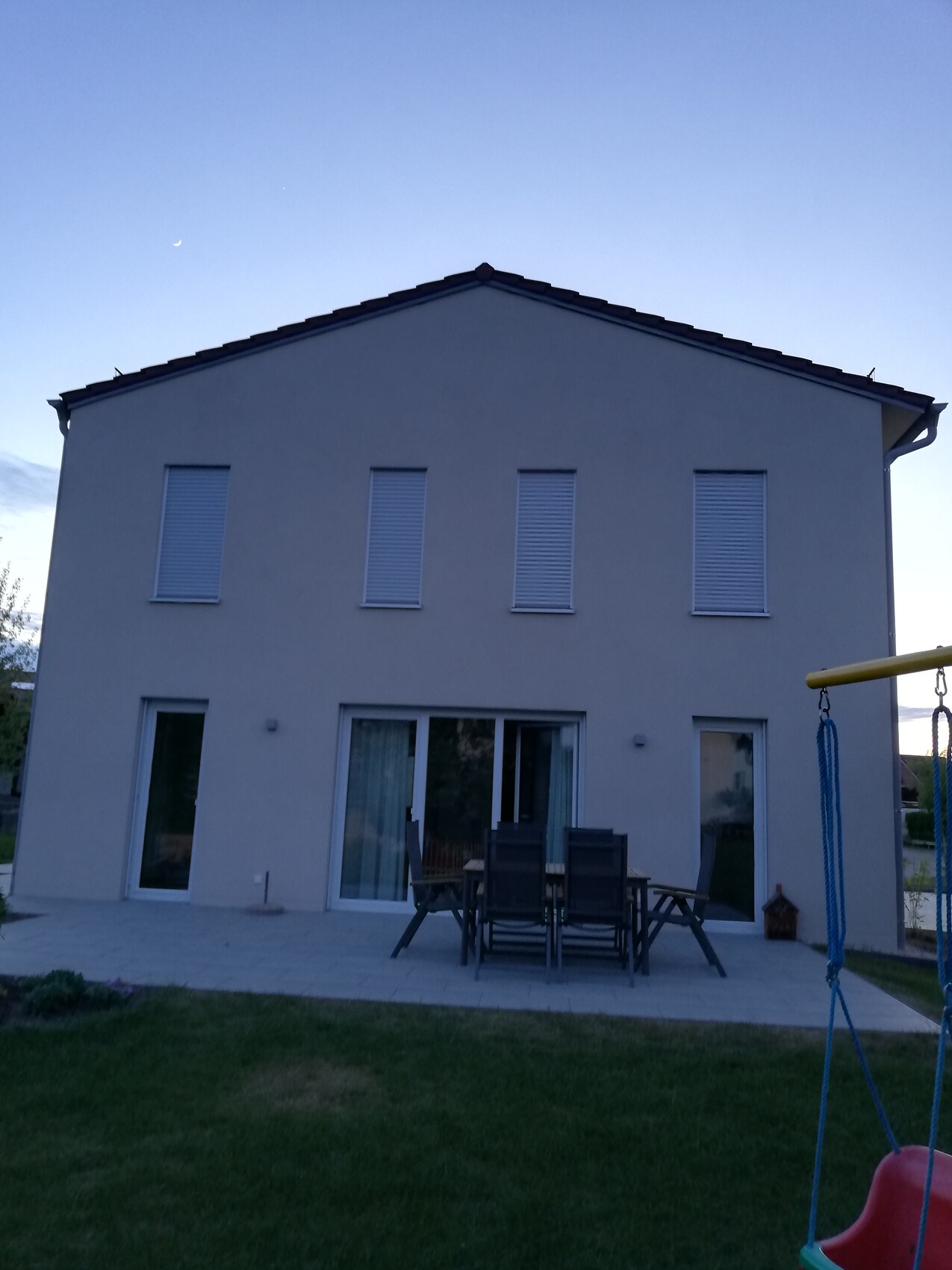 Two-story white house with terrace, dining table and chairs; play equipment in the garden.