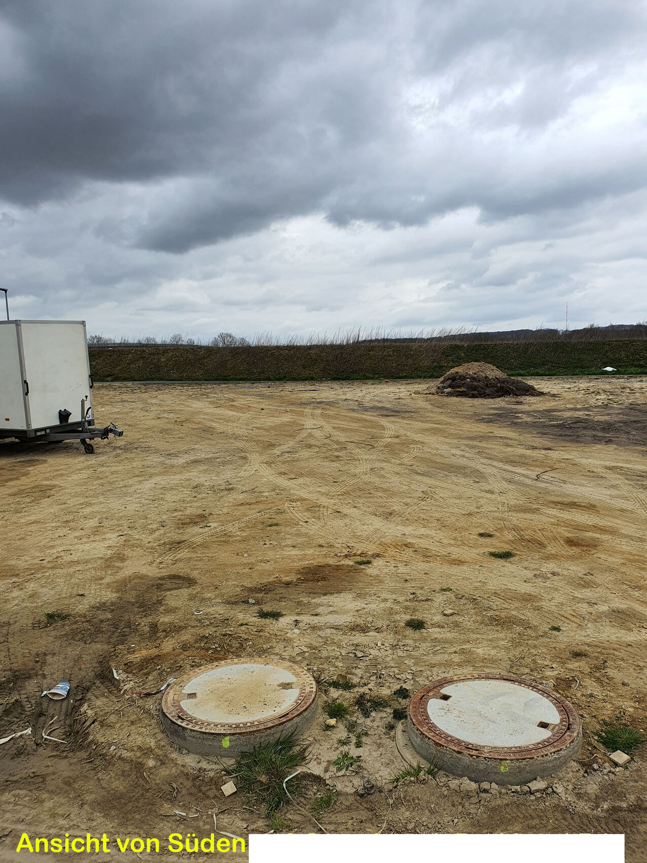 Construction site with two round manhole covers, trailer on the left, cloudy sky.