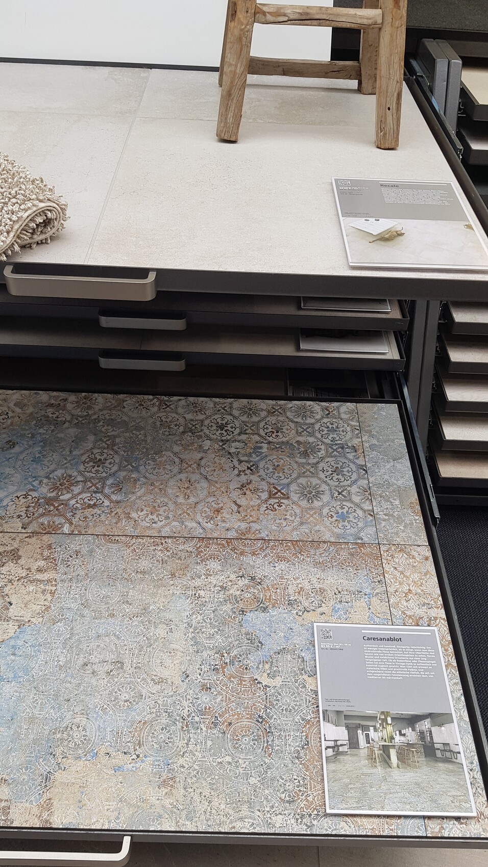 Showroom mit Fliesenmustern auf Regalen, Holzstuhl, Teppichrolle links und Infokarten.
