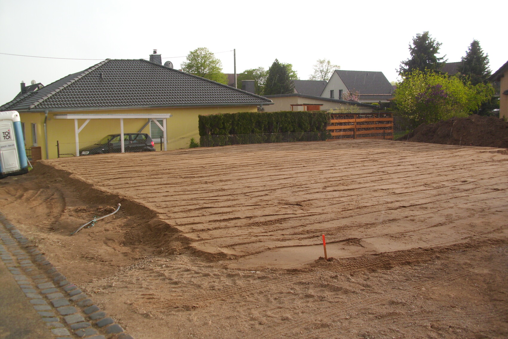 Baustelle mit ebener Erdfläche vor gelbem Einfamilienhaus mit Carport im Hintergrund.