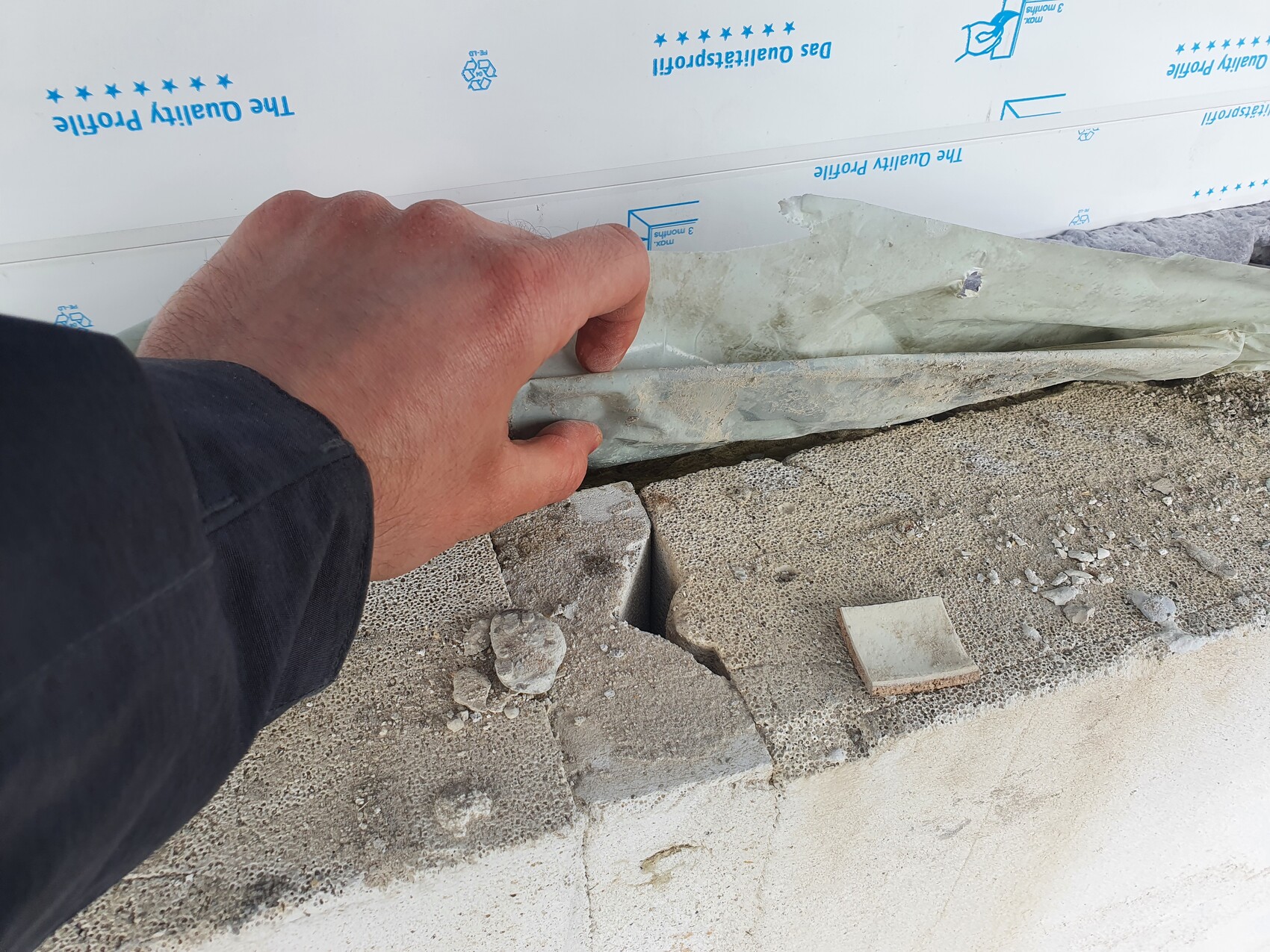 Close-up of a hand holding construction foil at a concrete wall joint; gravel and debris nearby.