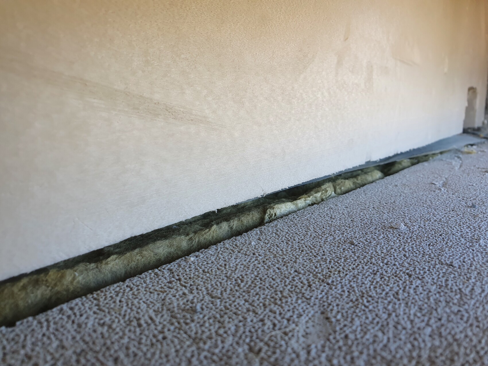 Bottom edge of a light-colored wall with a small gap to the floor slab and green insulation in the gap.