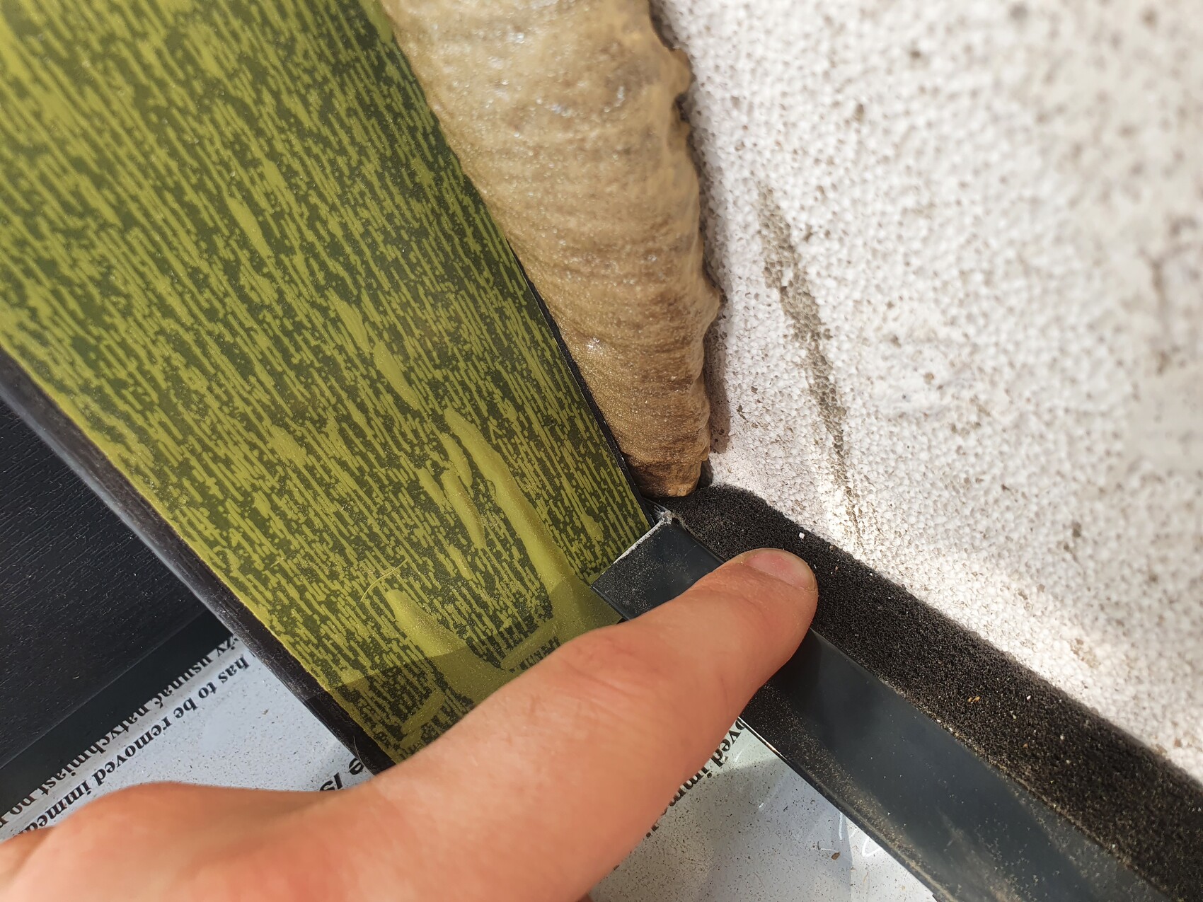 Hand pressing a black sealing strip into the joint between a green wooden panel and a rough wall.