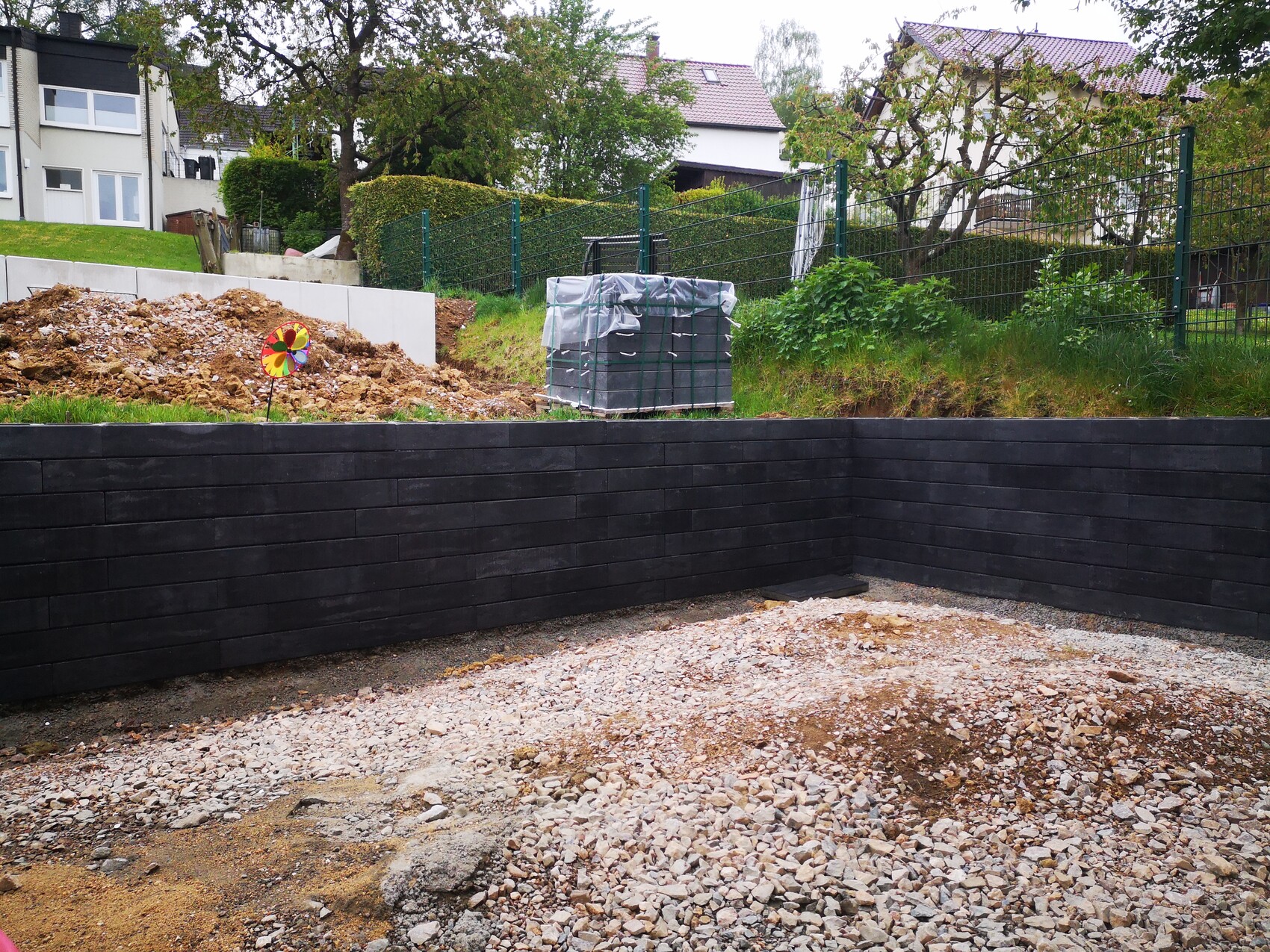 Construction site scene in the garden: black wooden panel wall, gravel ground, soil mound, colorful windmill.