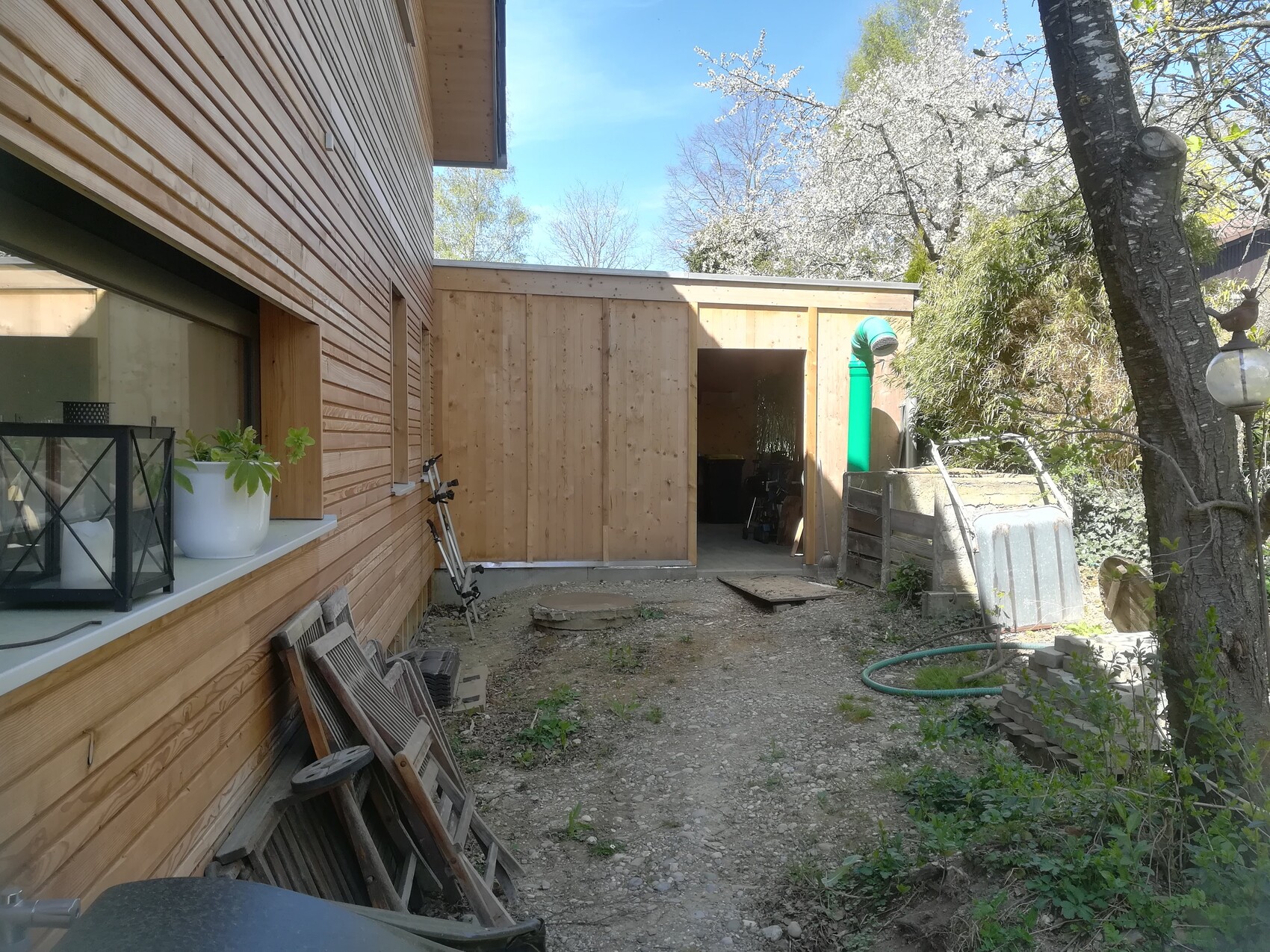 Exterior view of a wooden house with outbuilding, open entrance and garden tools.