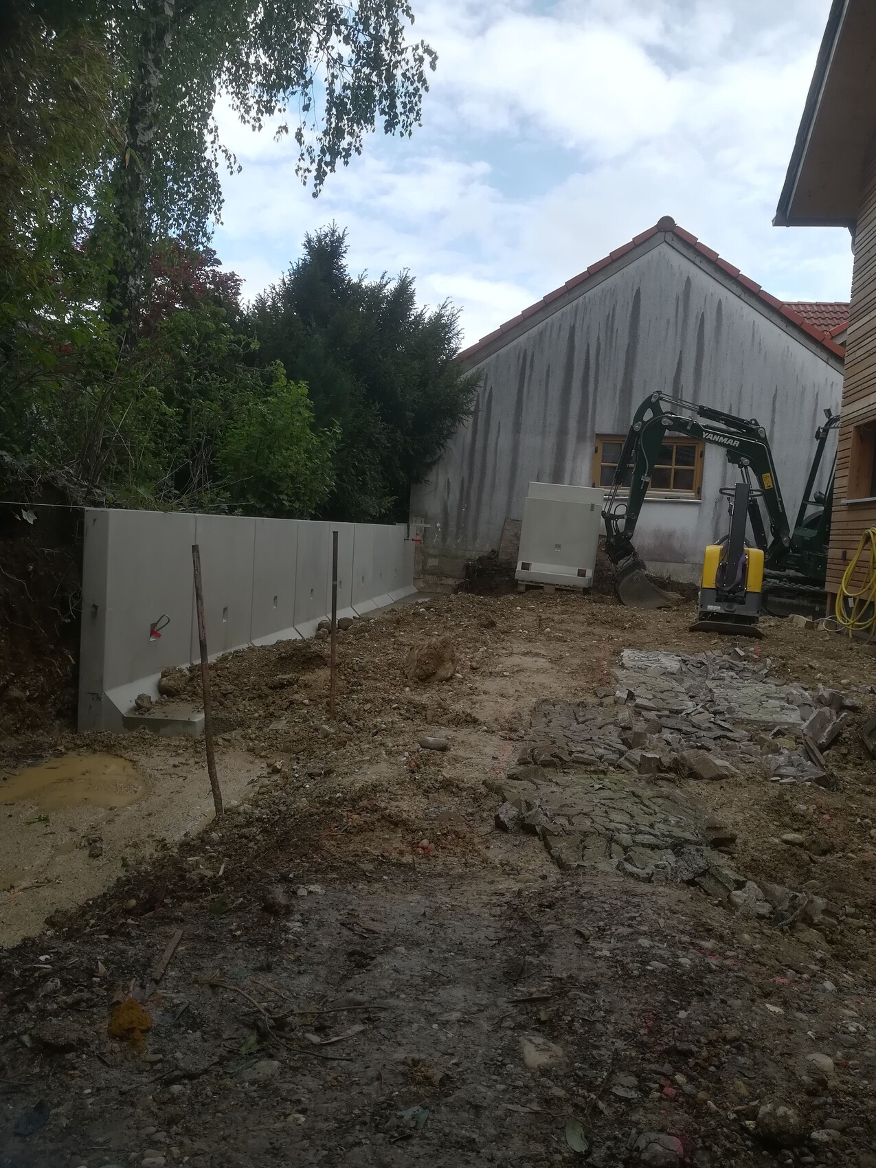 Small construction site with mini excavator next to house, concrete wall on the left, soil and rubble ground.