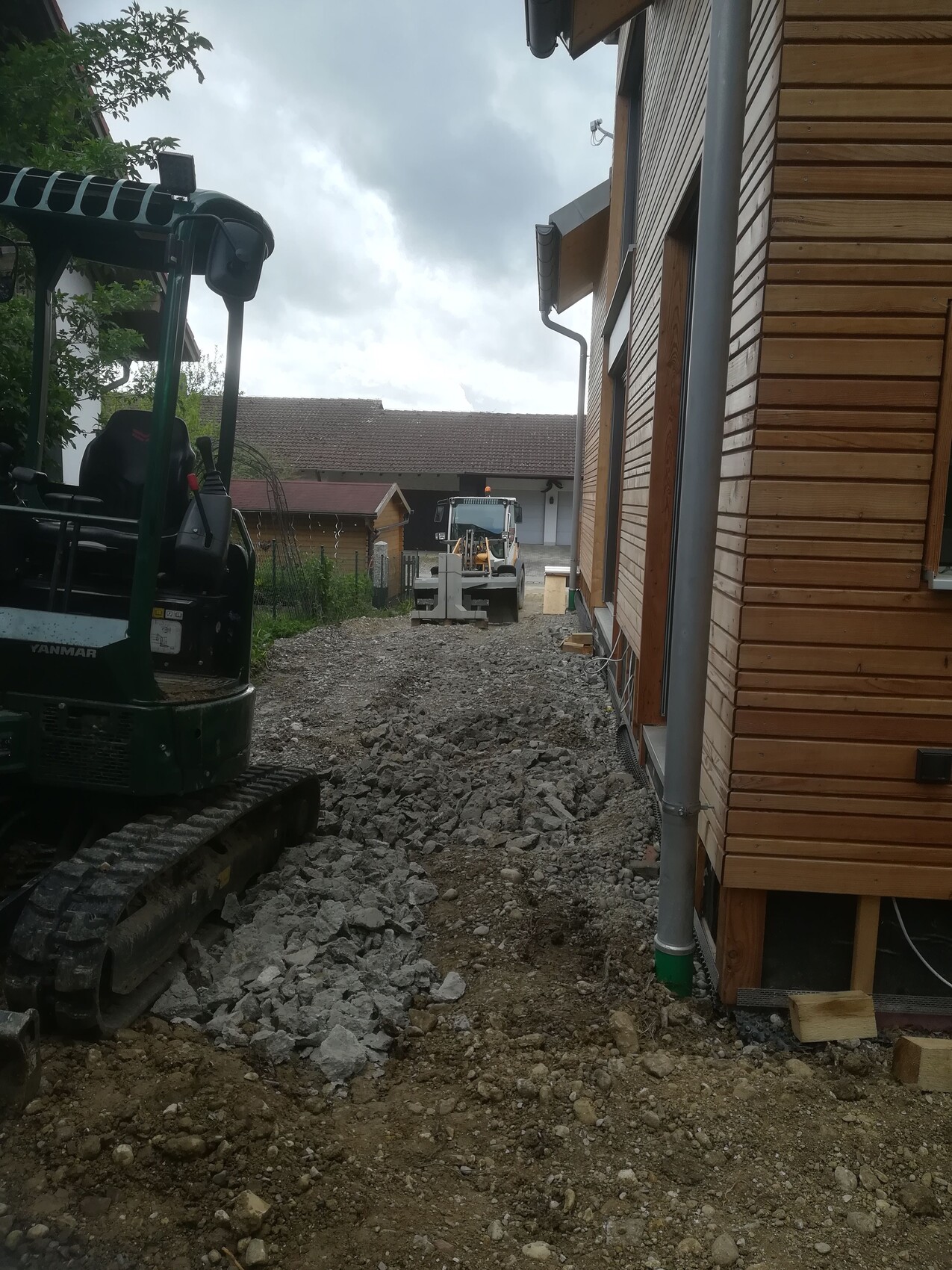 Construction site next to wooden house; unpaved gravel path, excavator on the left, construction equipment in the distance.