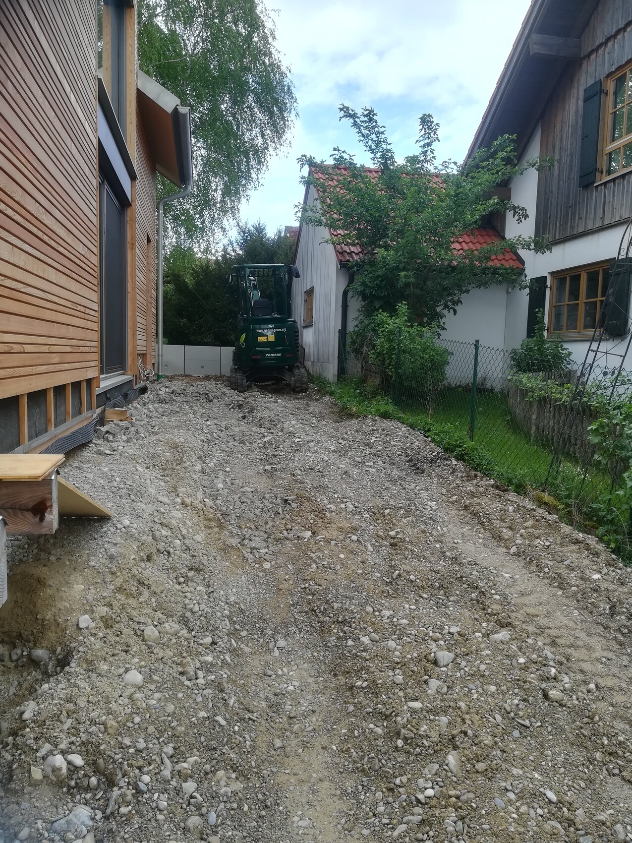 Small construction site: gravel path between wooden houses, mini excavator at the end of the property.
