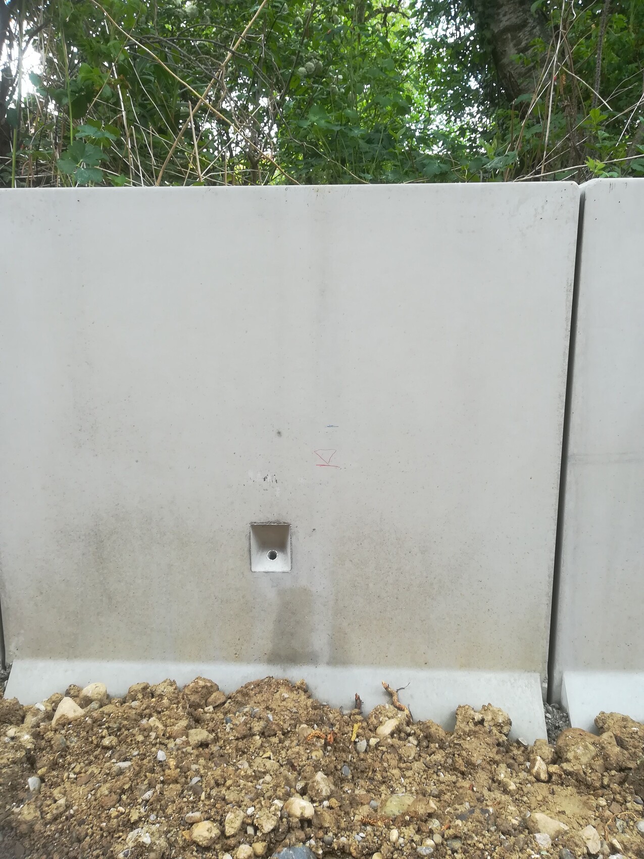 Gray concrete barrier with square metal fixture, gravel and soil in front, green background.