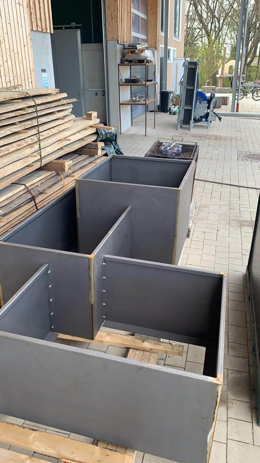 Several large gray metal containers are placed outdoors next to stacks of wooden planks.