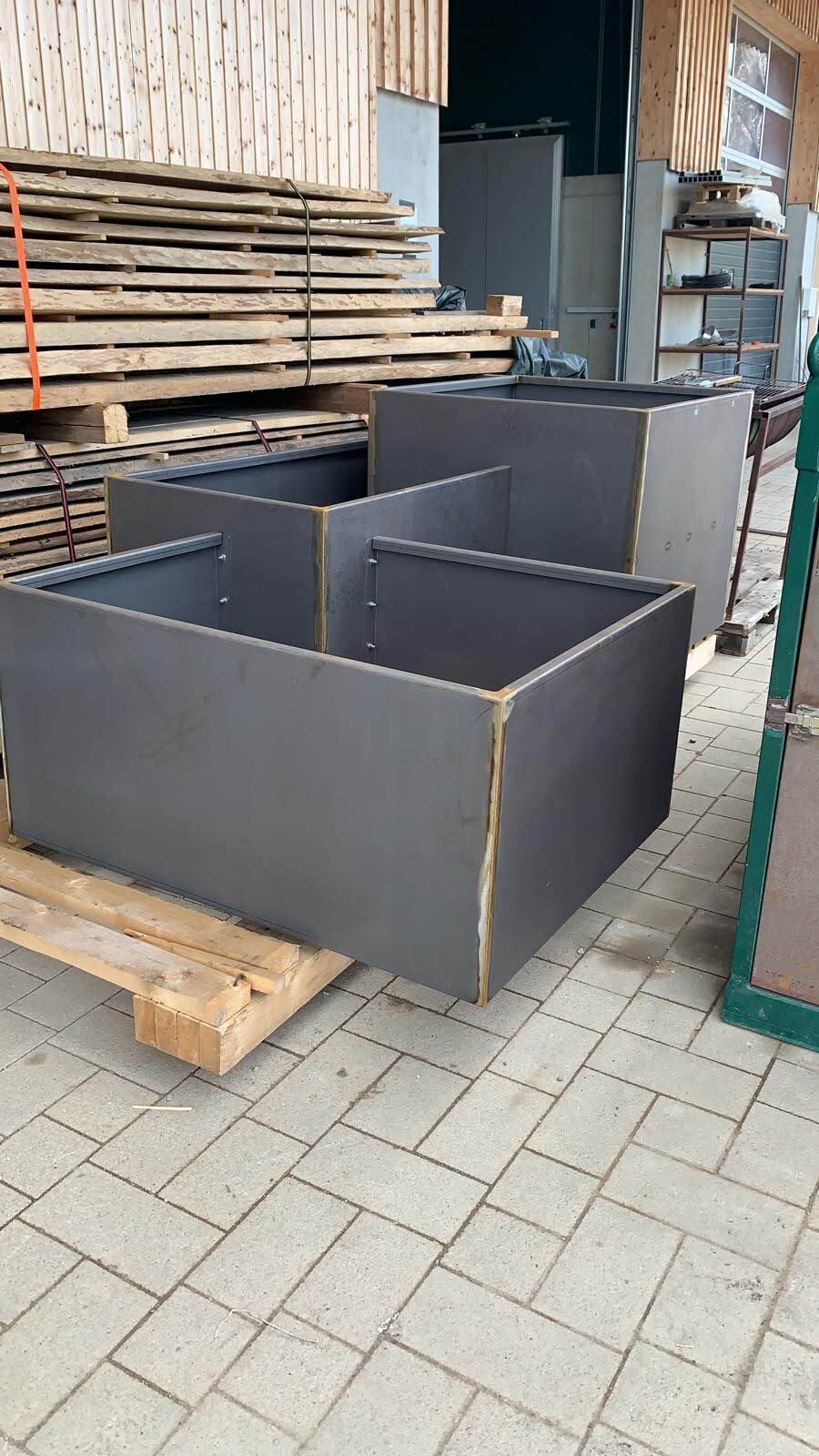 Several gray metal boxes stand outdoors next to stacked wood on pallets.