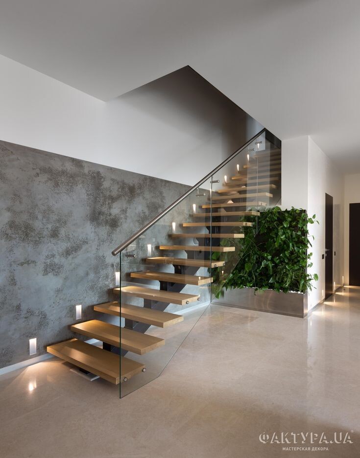 Modern staircase with wooden steps and glass railing, concrete wall on the left, plant wall on the right.