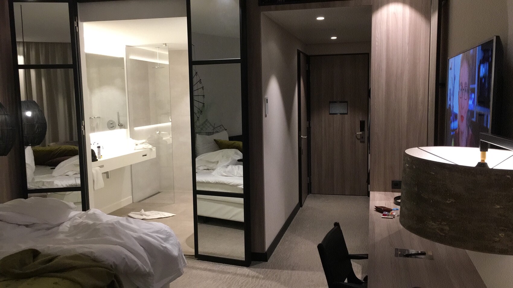 Hotel room with glass wall to the bathroom, unmade bed, TV and work area.